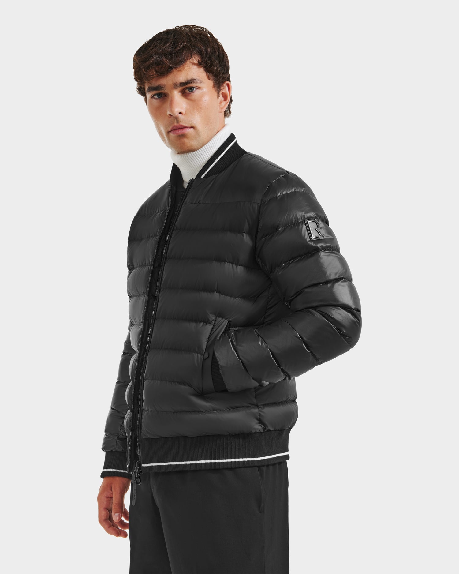 A man stands with his hands in his pockets wearing the Rudsak JEREMY - 6125025 Black men's down bomber jacket, paired with black pants and a white turtleneck, against a plain light background for a minimalist look.
