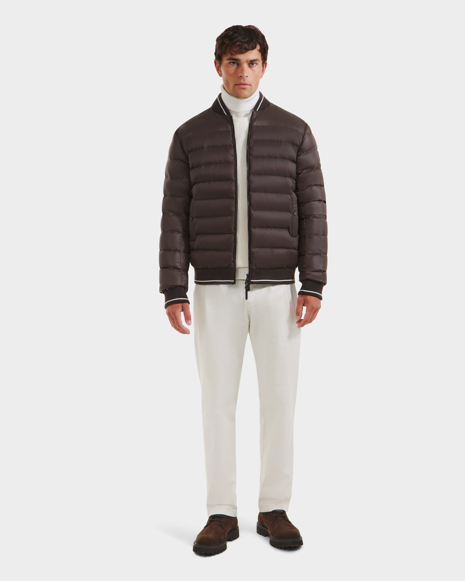 A man faces forward against a white background, wearing the Rudsak JEREMY - 6125025 Espresso men’s down bomber jacket over a white turtleneck and pants with dark brown shoes, showcasing minimalist style.