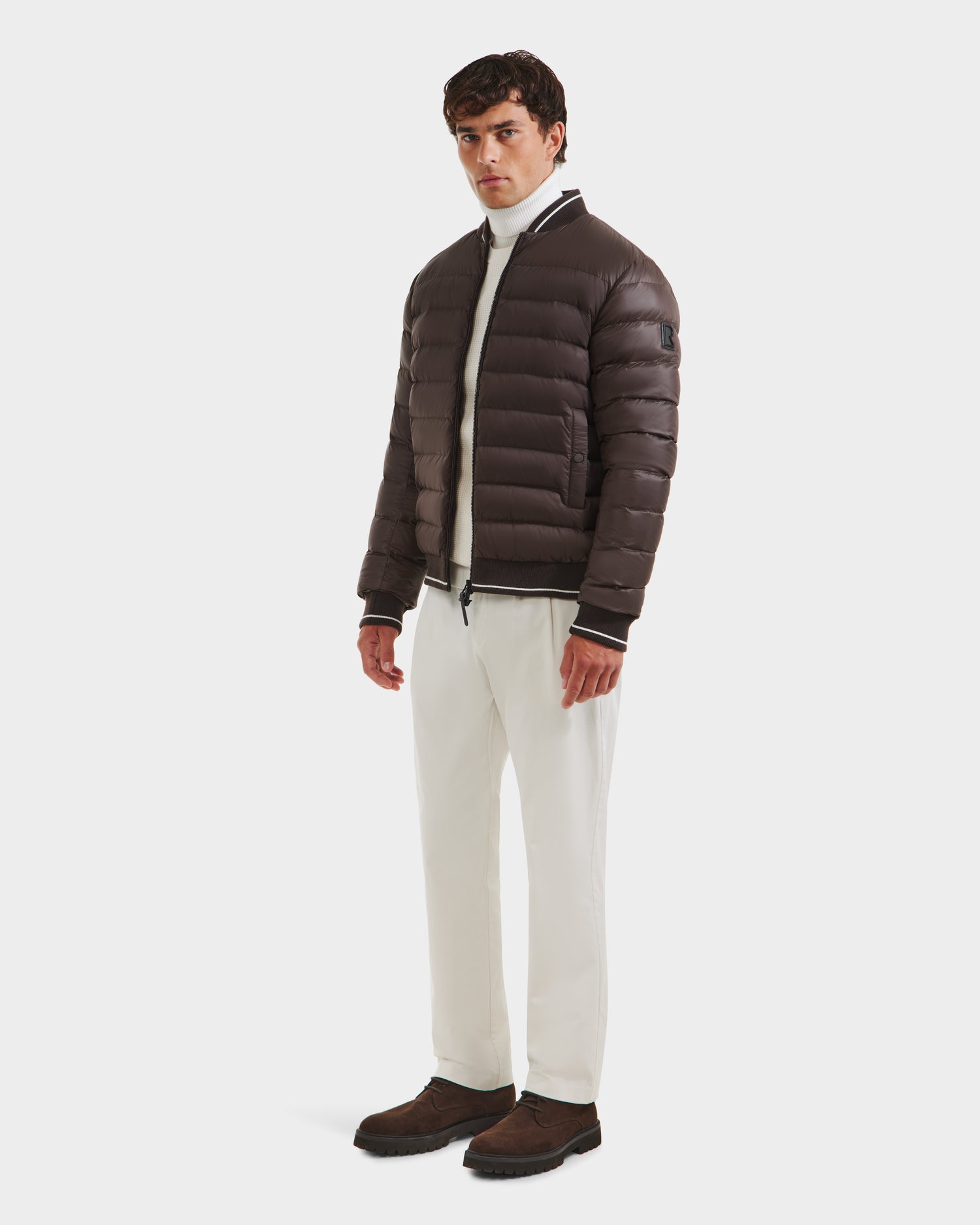 A man wears the Rudsak JEREMY - 6125025 Espresso premium puffer jacket over a white turtleneck, white pants, and brown shoes against a plain white background—ideal for minimalist wardrobes.
