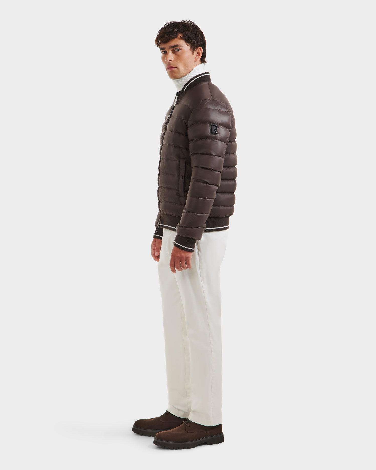 A man stands sideways wearing the Rudsak JEREMY - 6125025 Espresso men's down bomber jacket, paired with white pants and dark brown shoes, ideal for minimalist wardrobes, against a plain white background.