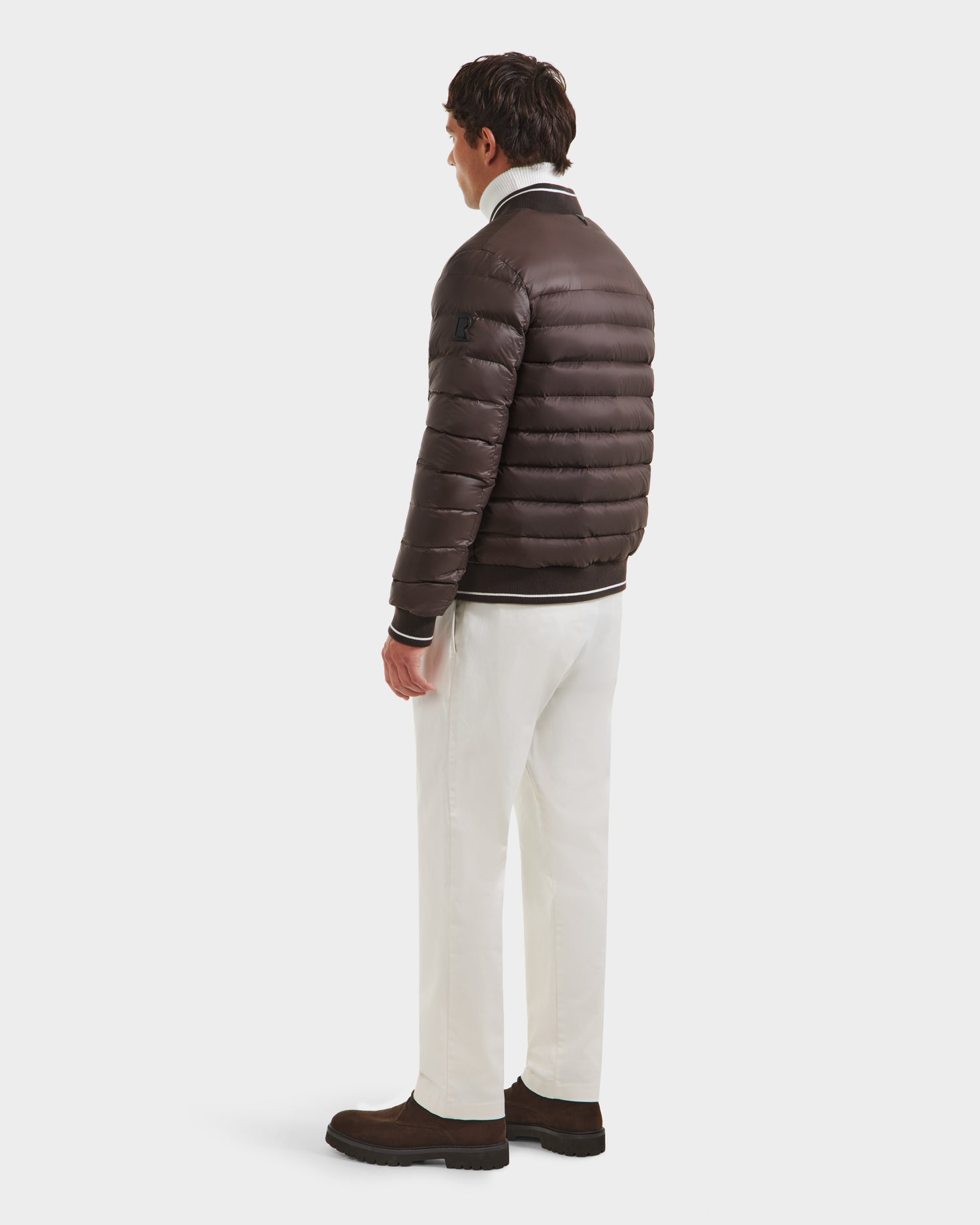 A man stands facing slightly away, wearing the Rudsak JEREMY - 6125025 Espresso men’s down bomber jacket with white pants and brown boots, creating a minimalist look against a plain white background.