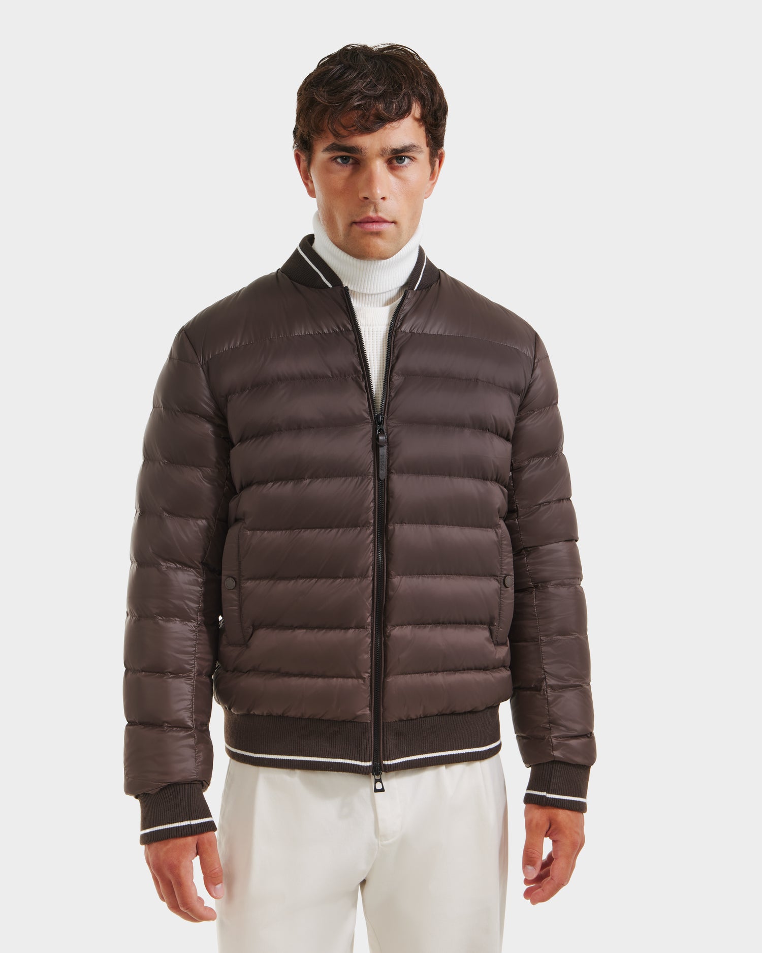 A man wearing the Rudsak JEREMY - 6125025 Espresso down bomber jacket, a white turtleneck sweater, and white pants stands against a plain white background.