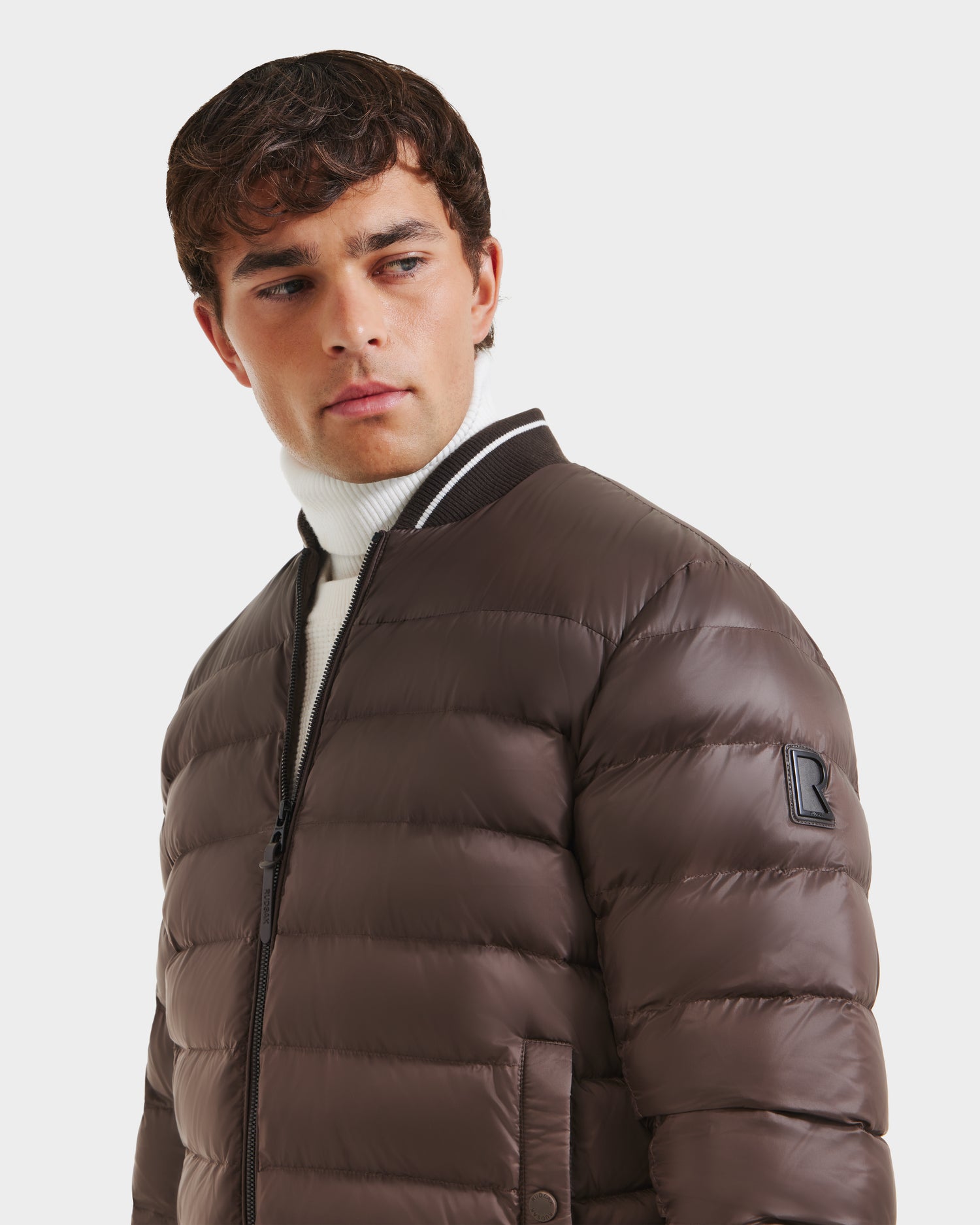 A young man looks to the side wearing the Rudsak JEREMY - 6125025 Espresso men’s down bomber with premium insulation, black collar, and sleeve patch over a white turtleneck, set against a plain white background.