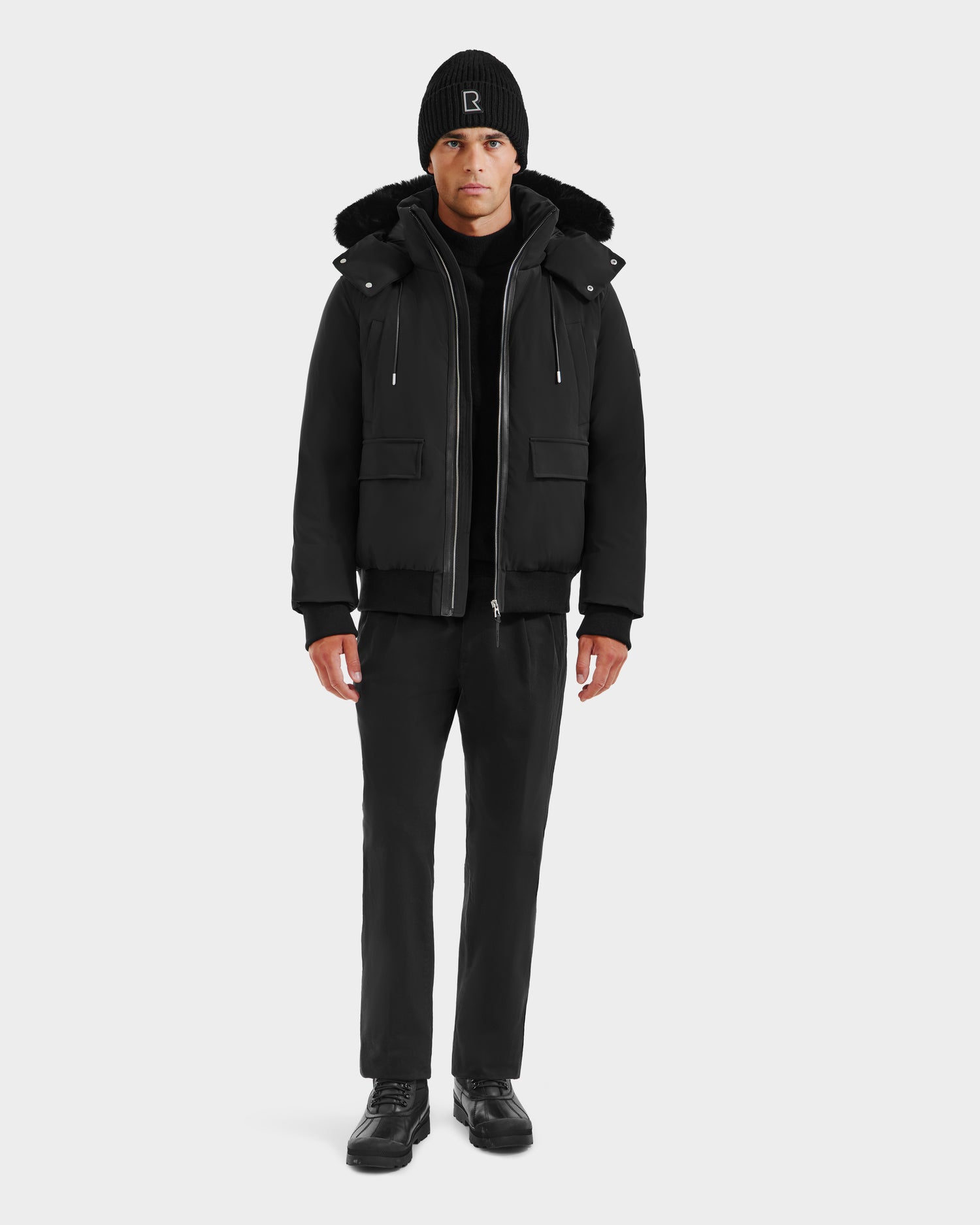 A man stands facing forward in the Rudsak JEROME - 6125514 Black men’s down bomber jacket with RDS-certified down, styled with a black beanie, pants, and shoes against a light background.