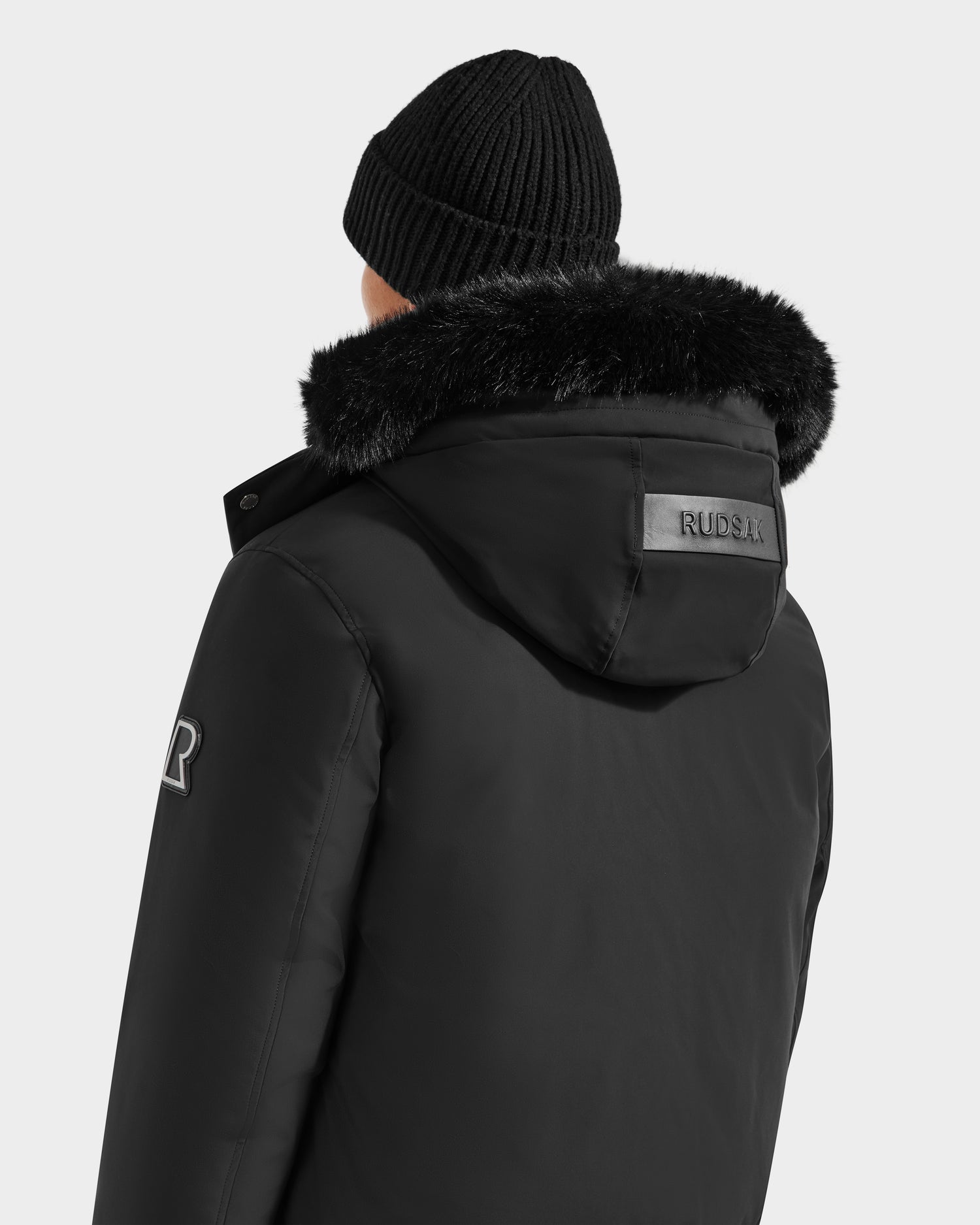 A person shown from behind wears the Rudsak JEROME - 6125514 Black knit beanie and winter jacket with faux fur-lined hood, RUDSAK branding on the hood and sleeve patch, filled with RDS-certified down for warmth.