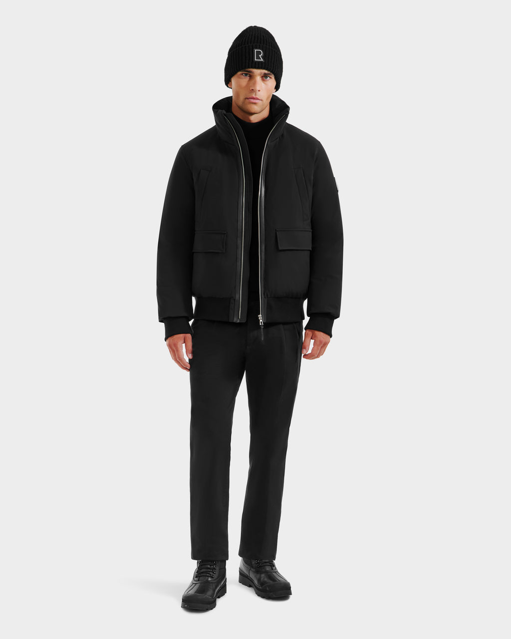 A man faces forward against a plain light background, wearing the Rudsak JEROME - 6125514 Black men’s down bomber jacket, paired with a black beanie, pants, and shoes.
