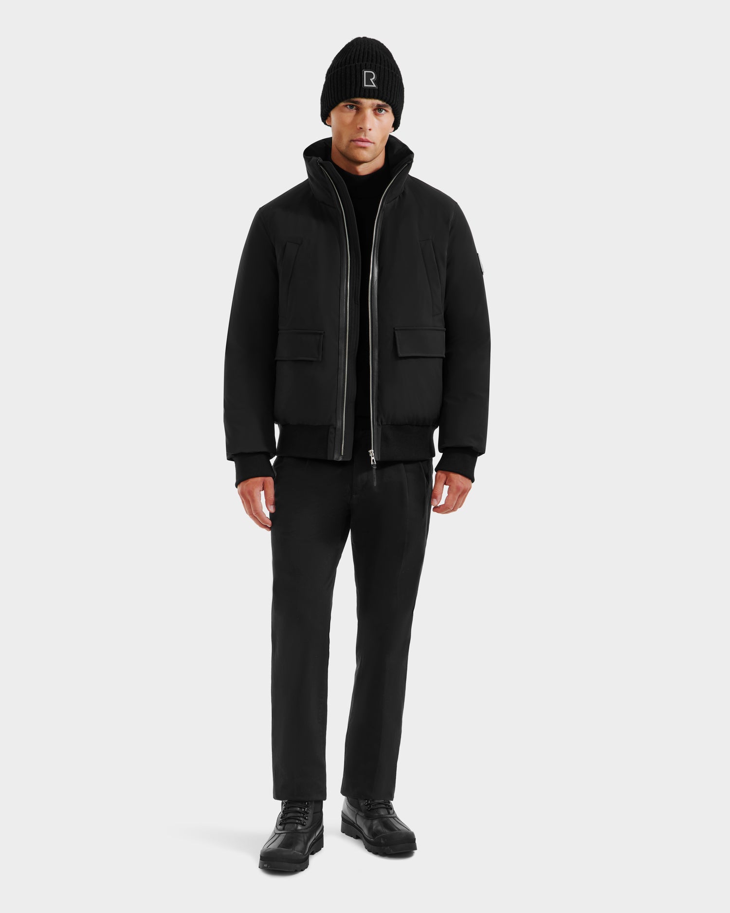 A man faces forward against a plain light background, wearing the Rudsak JEROME - 6125514 Black men’s down bomber jacket, paired with a black beanie, pants, and shoes.