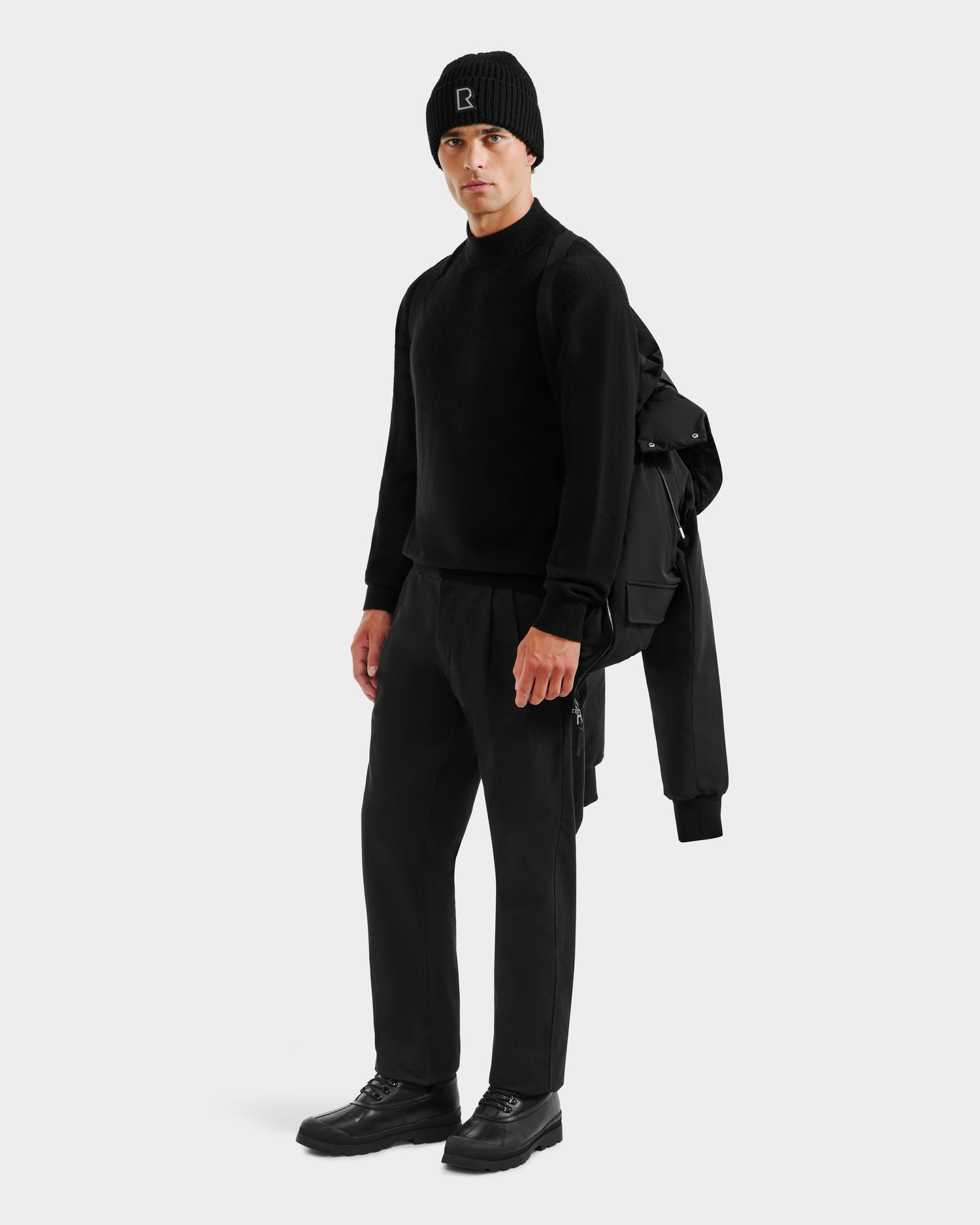 A person stands facing forward, wearing all-black attire with the Rudsak JEROME - 6125514 Black men’s down bomber jacket draped over one shoulder. The RDS-certified down offers warmth and style. They pose against a plain white background.