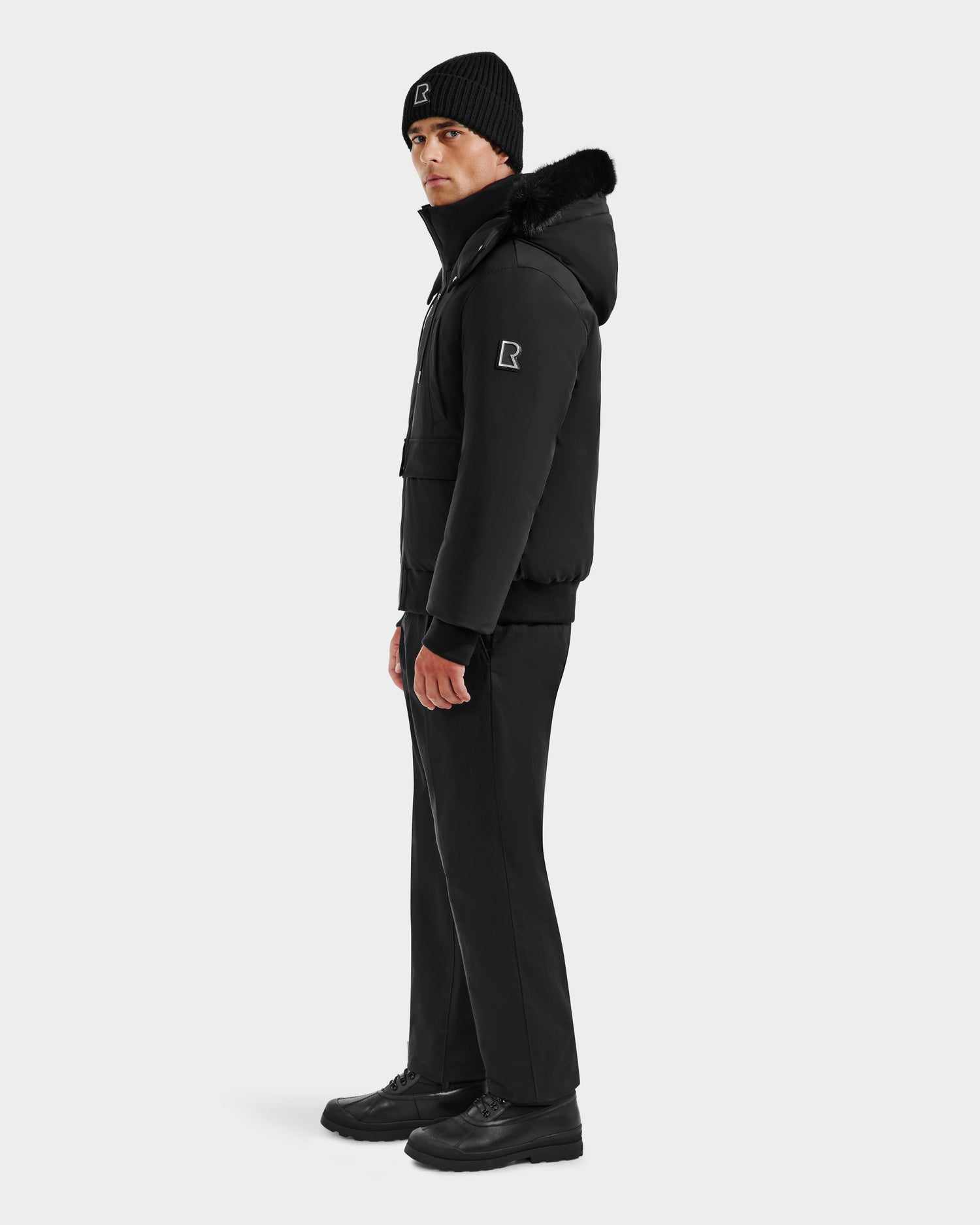 A man stands sideways against a plain background, wearing Rudsak’s JEROME - 6125514 Black men’s down bomber jacket with fur trim, black pants, and shoes—all featuring the R logo—highlighting its winter performance style.
