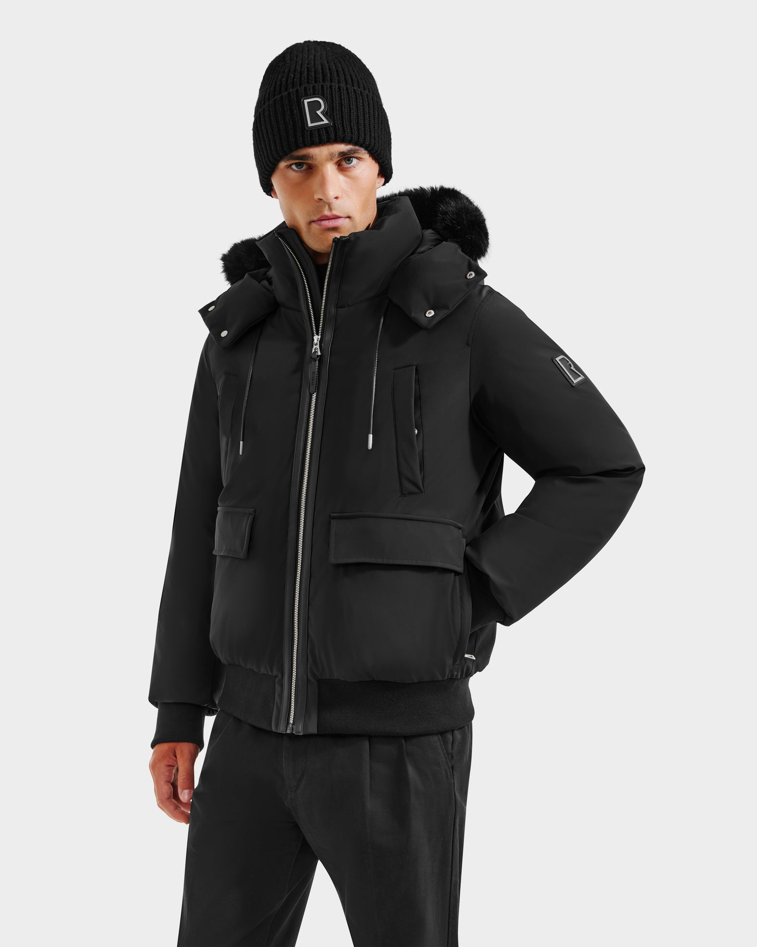 A man wears the Rudsak JEROME - 6125514 Black men’s down bomber jacket with fur trim and a black beanie, standing with one hand in his pocket against a plain light background.