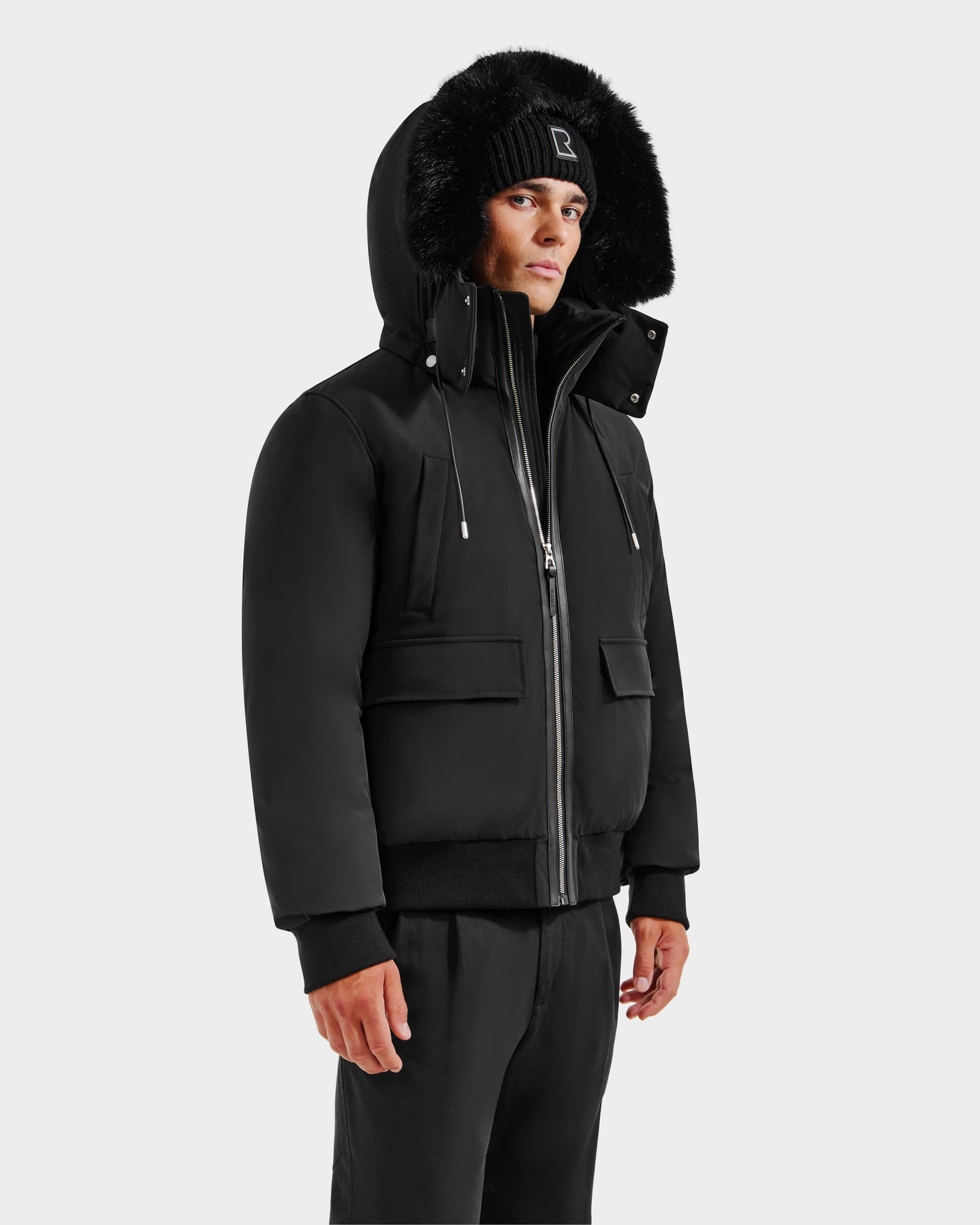 A person wears the Rudsak JEROME - 6125514 Black men’s down bomber with a fur-lined hood, styled with a black knit beanie and black pants, standing against a plain white background.