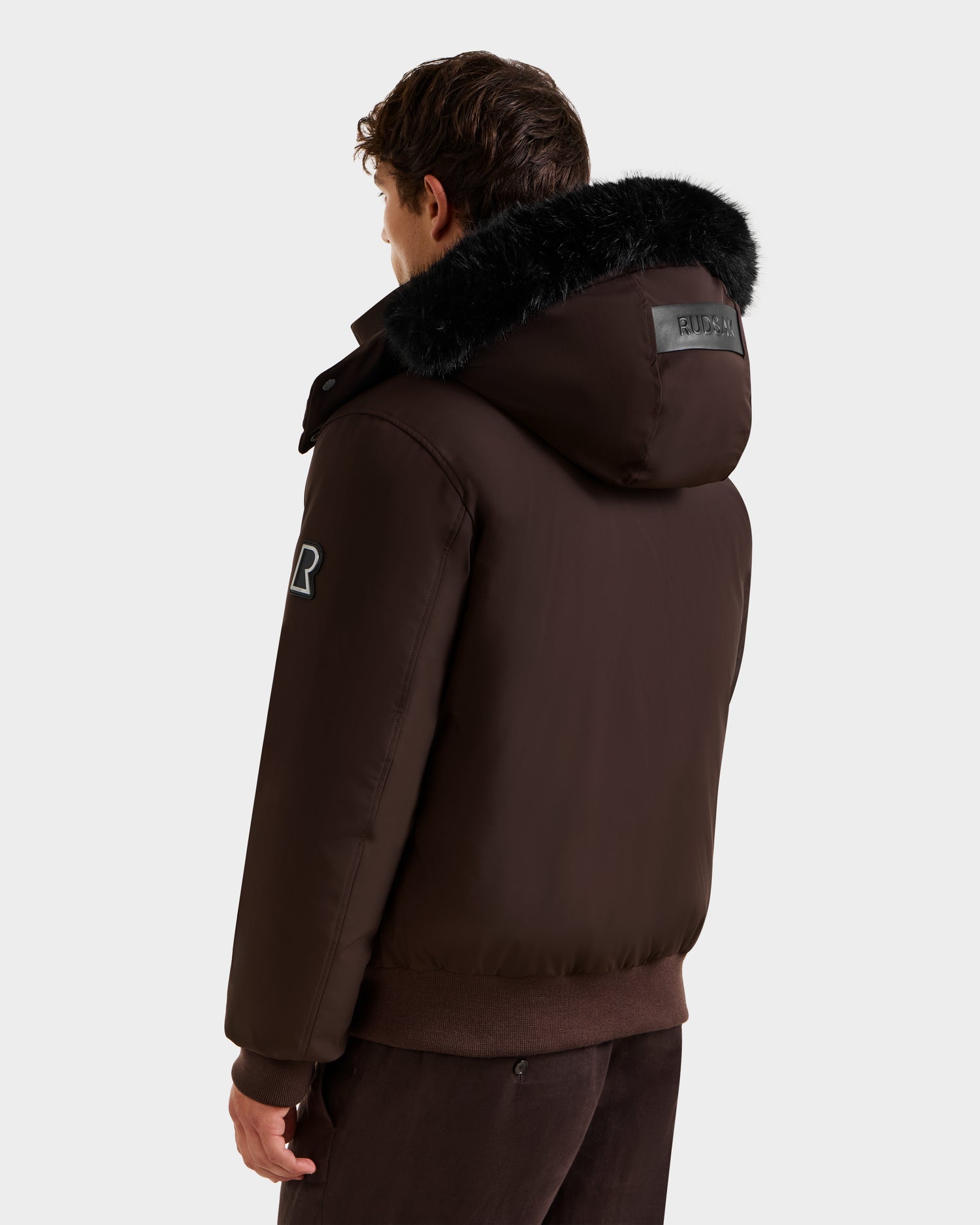 A person wearing the Rudsak JEROME - 6125514 Espresso men’s down bomber jacket with black fur trim on the hood is seen from behind against a plain white background.