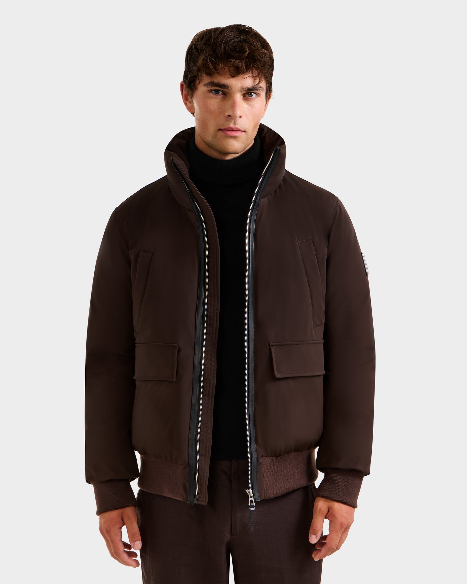 A man wears the Rudsak JEROME - 6125514 Espresso, a dark brown RDS-certified down bomber jacket with a high collar, zip closure, and front pockets over a black turtleneck against a plain light background.