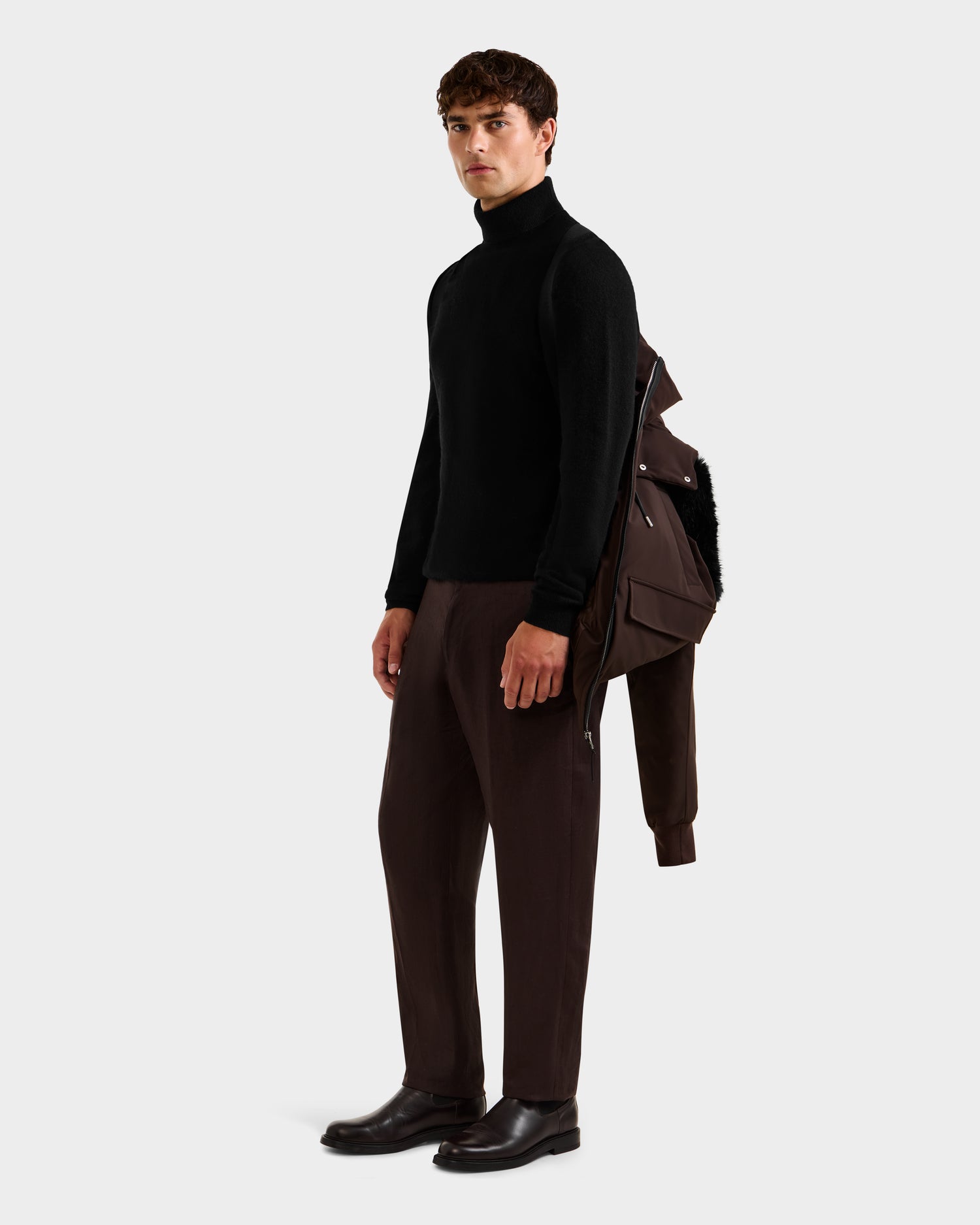 A man stands against a plain background, wearing a black turtleneck sweater, dark brown pants, black shoes, and the Rudsak JEROME - 6125514 Espresso men’s down bomber jacket draped over one shoulder.