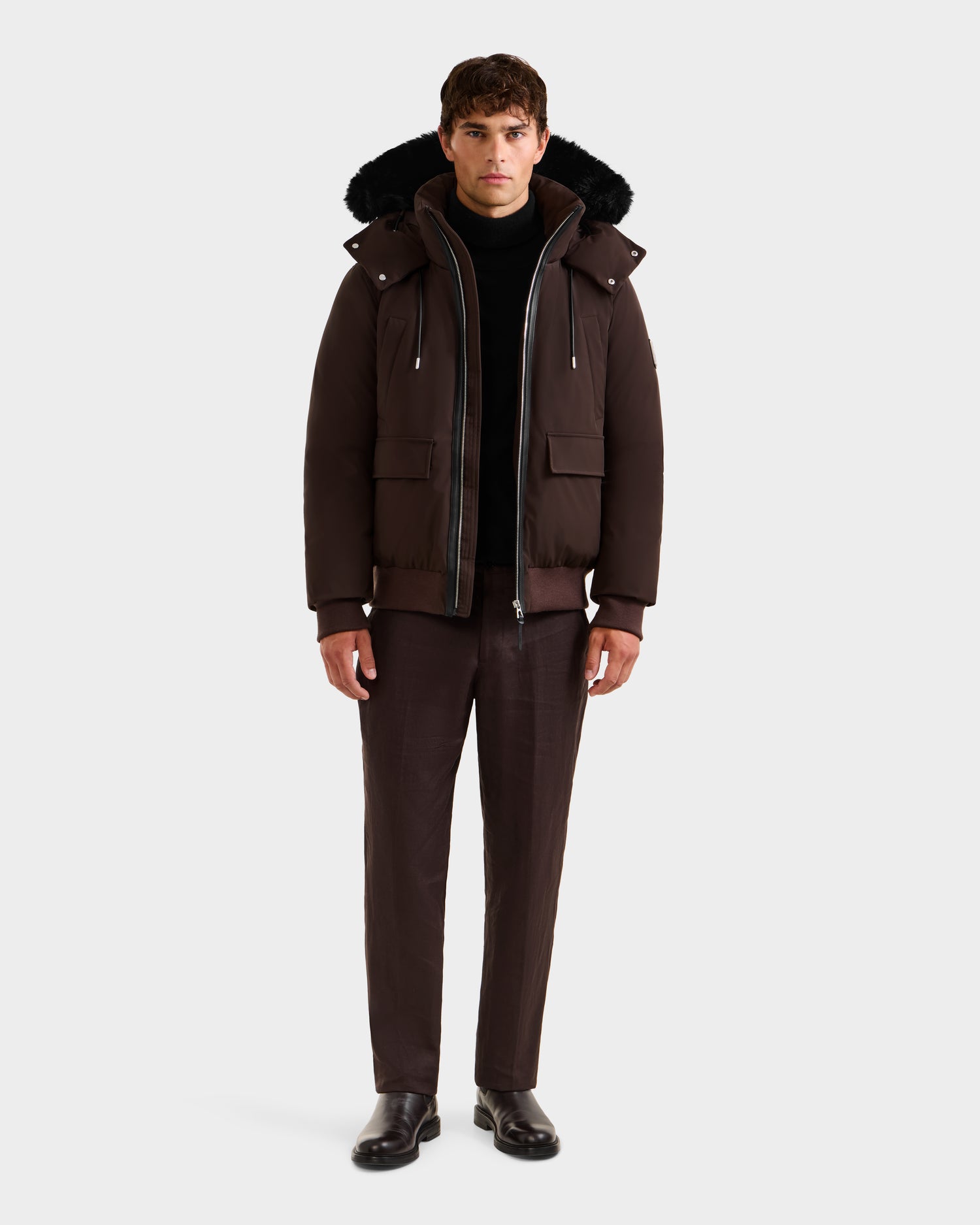 A man stands facing forward against a plain white background, wearing the Rudsak JEROME - 6125514 Espresso winter performance jacket with hood, paired with a black turtleneck, brown trousers, and black shoes.
