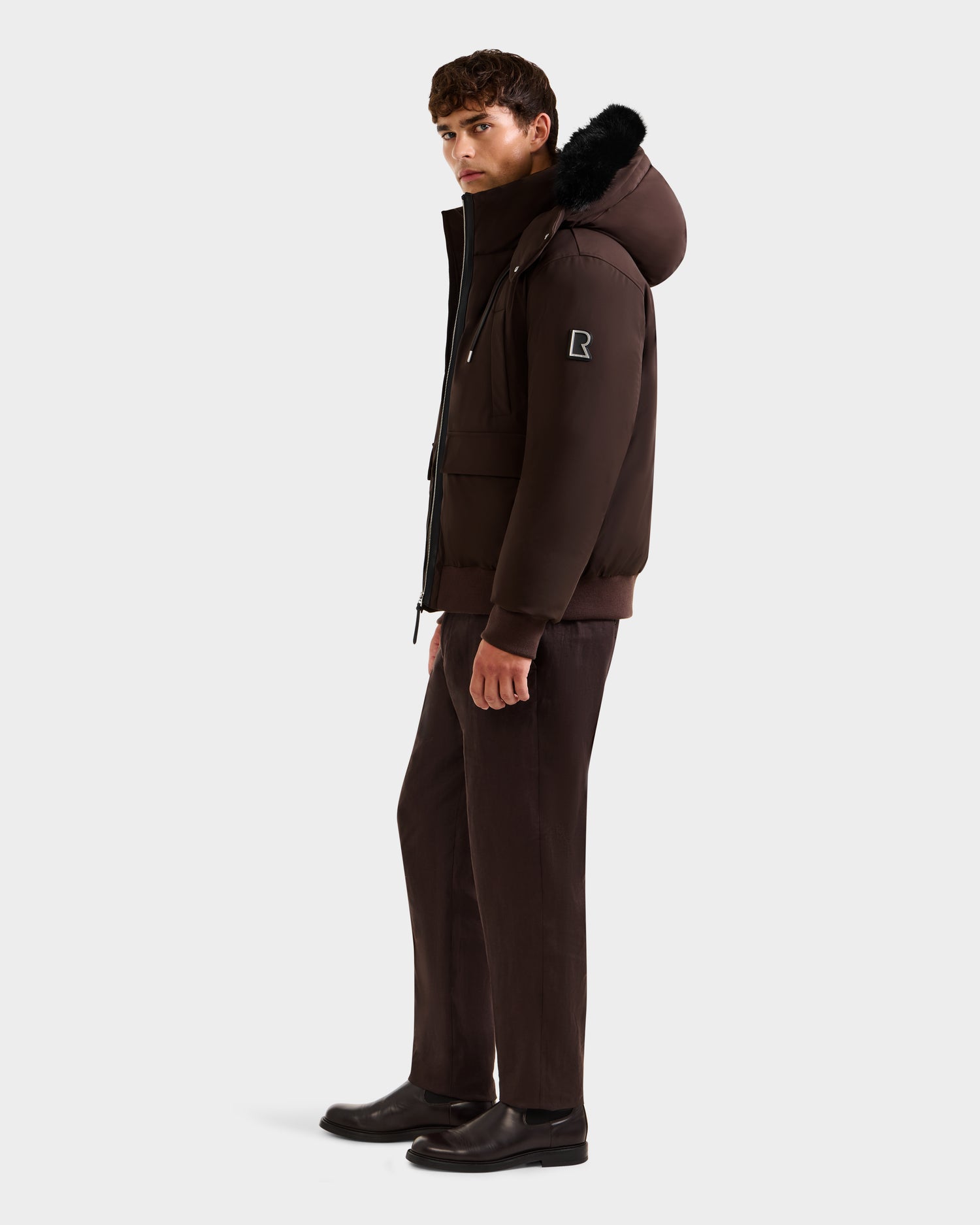 A man stands in profile against a white background, wearing the Rudsak JEROME - 6125514 Espresso mens down bomber jacket with black fur trim, paired with matching brown pants and black shoes.
