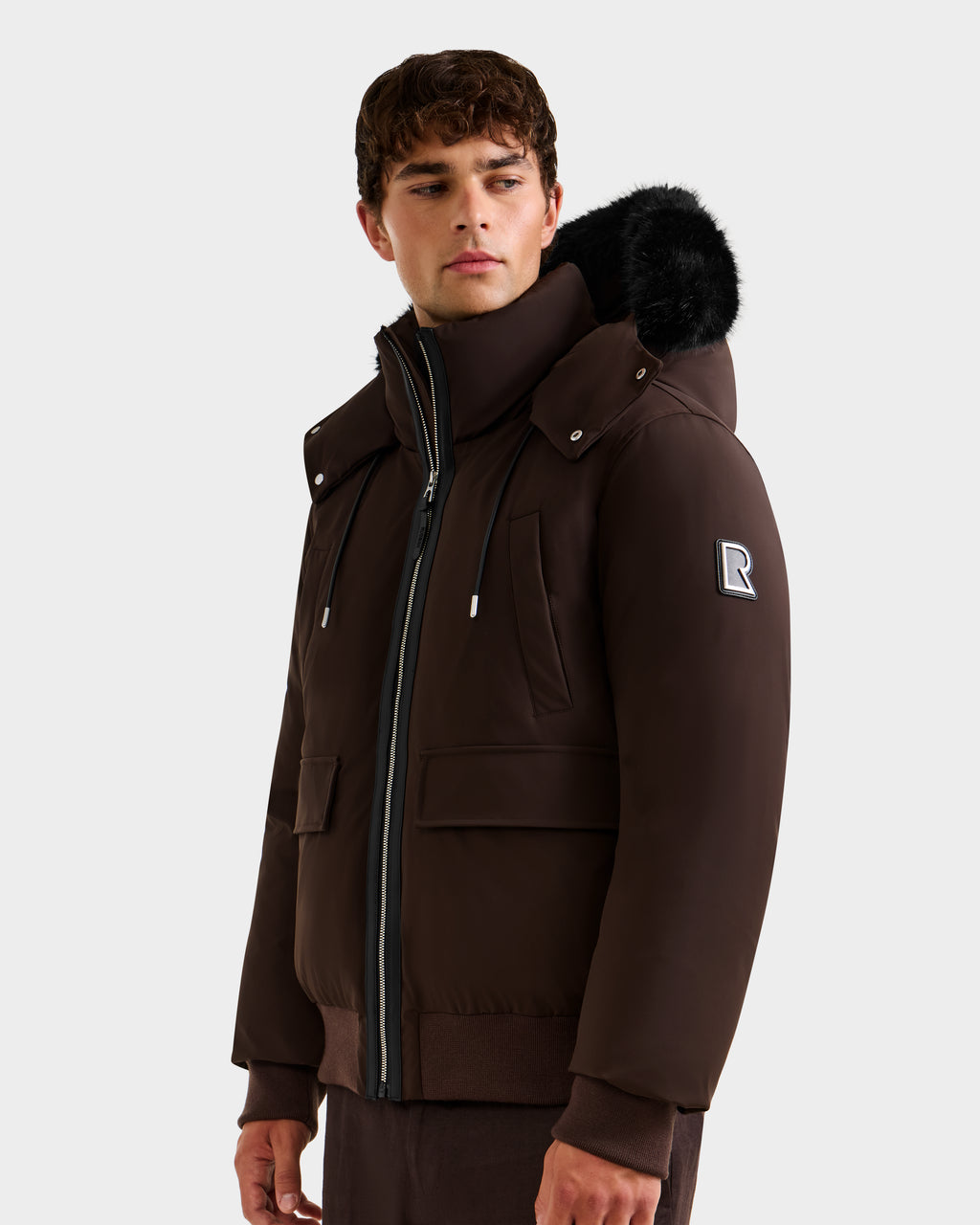 A young man stands against a plain background, wearing the Rudsak JEROME - 6125514 Espresso men’s down bomber jacket with a fur-lined hood, zip-up front, winter performance features, and an "R" patch on the sleeve.