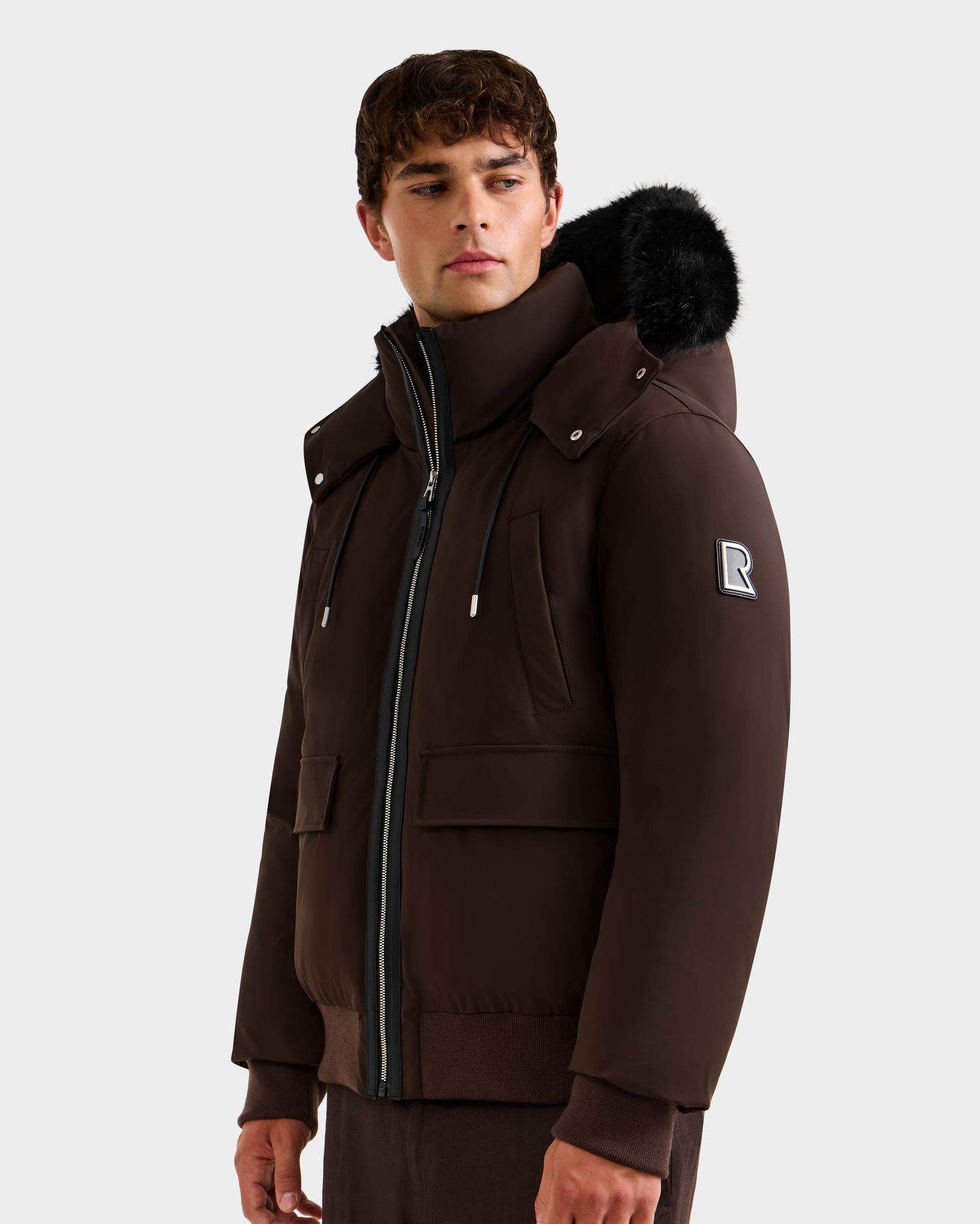 A young man stands against a plain background, wearing the Rudsak JEROME - 6125514 Espresso men’s down bomber jacket with a fur-lined hood, zip-up front, winter performance features, and an "R" patch on the sleeve.
