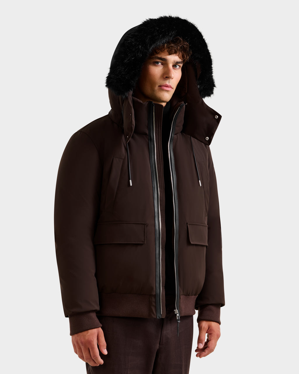 A man models the Rudsak JEROME - 6125514 Espresso, a dark brown mens down bomber jacket with black fur lining, double front zippers, flap pockets, and matching pants against a plain light background.