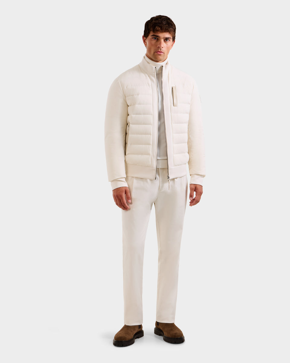 A man faces the camera in a sleek, all-white outfit featuring the Rudsak JIMMY - 6125723 Vanilla men’s quilted down jacket, made with RDS-certified down and water- and wind-resistant, paired with brown shoes against a plain background.