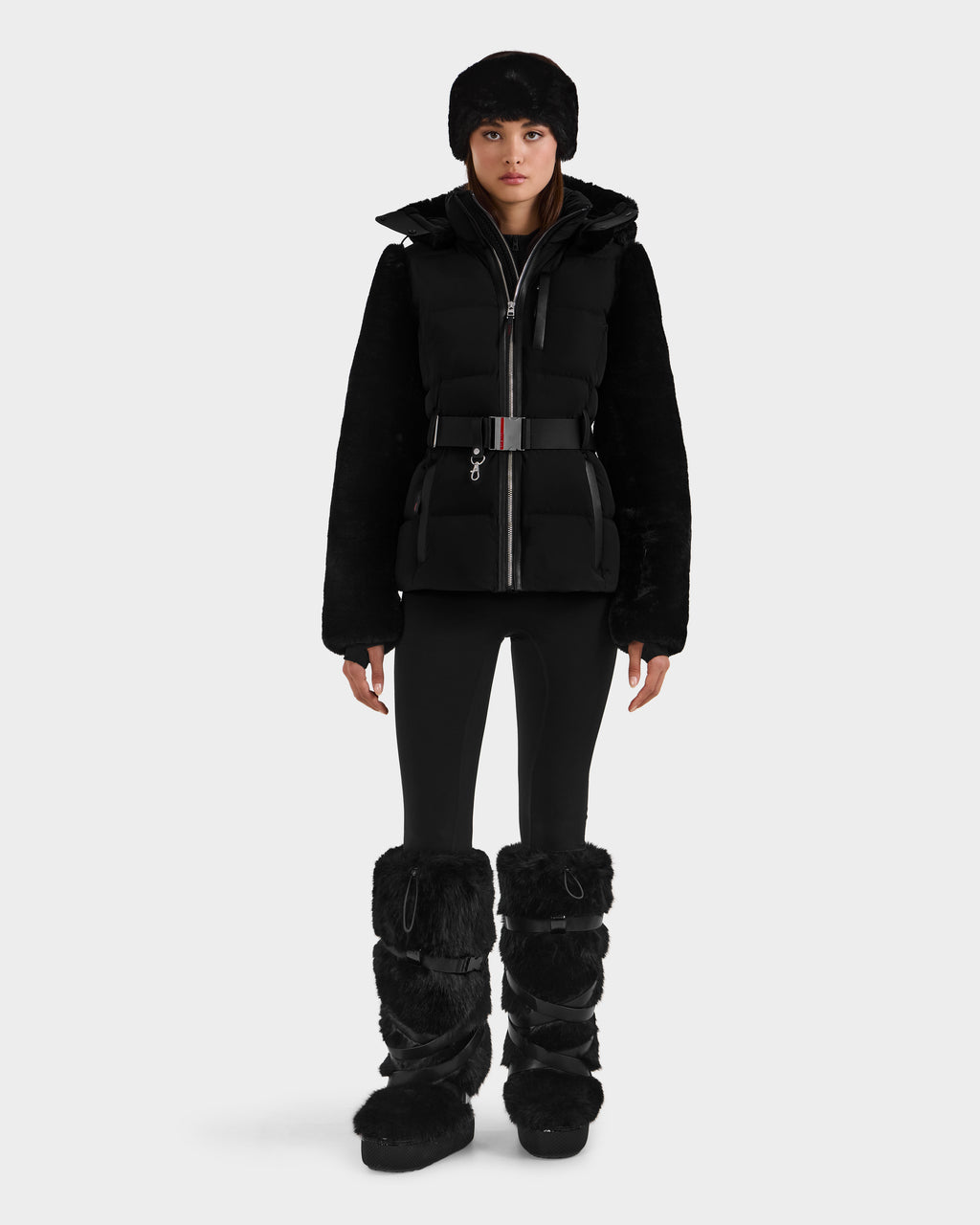 A person wears the Rudsak JOANNA F - 8125593 Black puffer jacket with belted waist, paired with fitted pants, a furry hat, and knee-high black furry boots. Pre-order now—ships by November 30th.