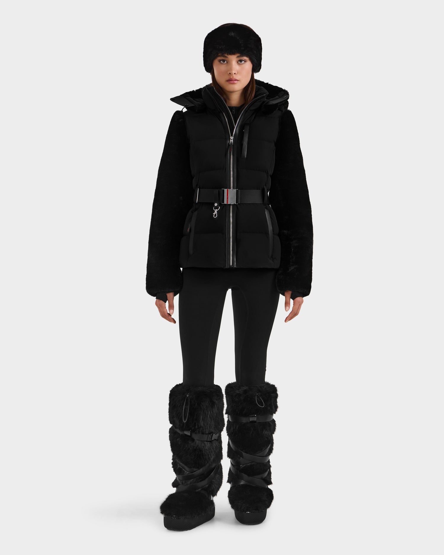 A person wears the Rudsak JOANNA F - 8125593 Black puffer jacket with belted waist, paired with fitted pants, a furry hat, and knee-high black furry boots. Pre-order now—ships by November 30th.