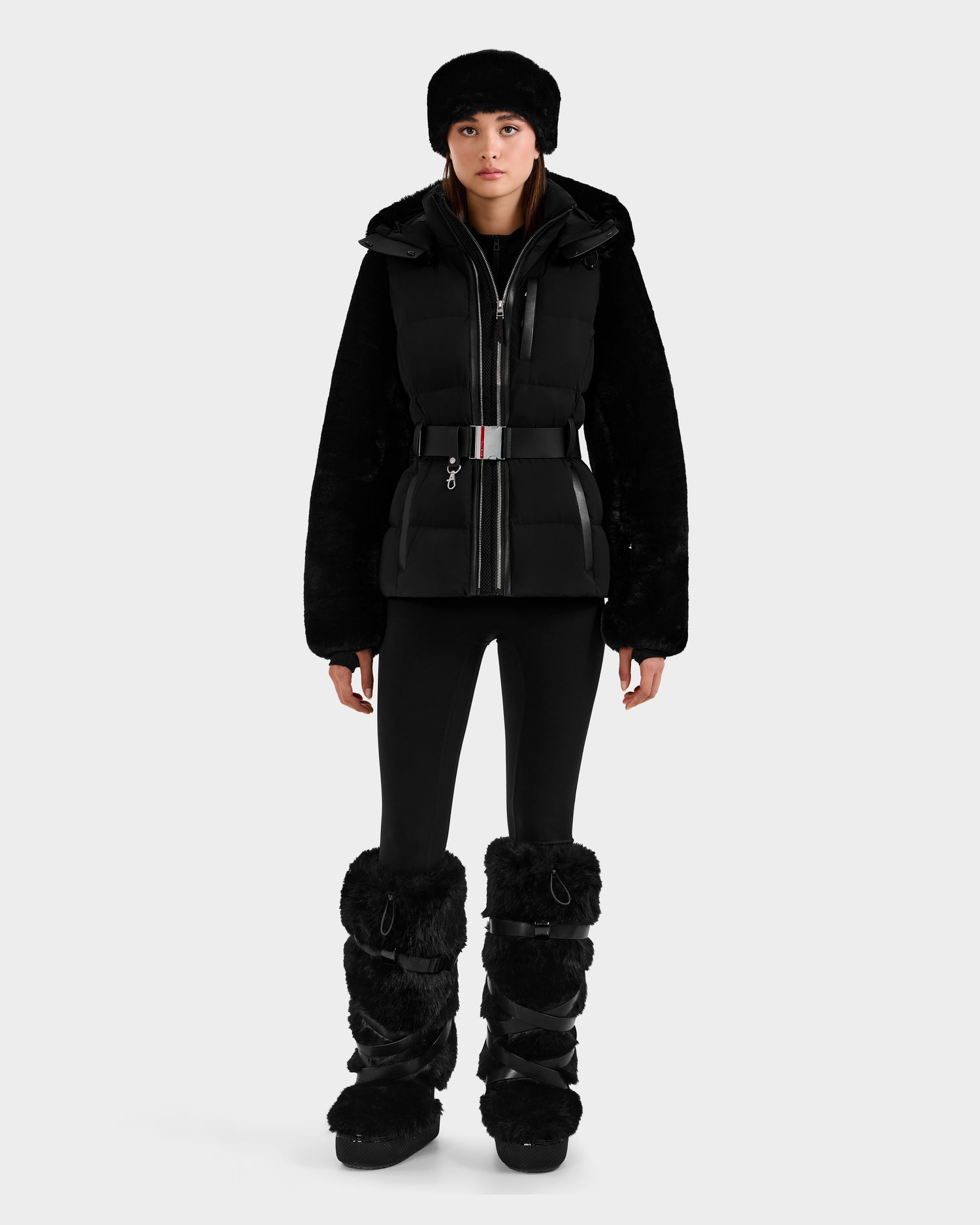 A person wears the Rudsak JOANNA F - 8125593 Black women’s down ski jacket with RECCO rescue technology, a fur hat, leggings, and furry boots, standing against a plain white background.