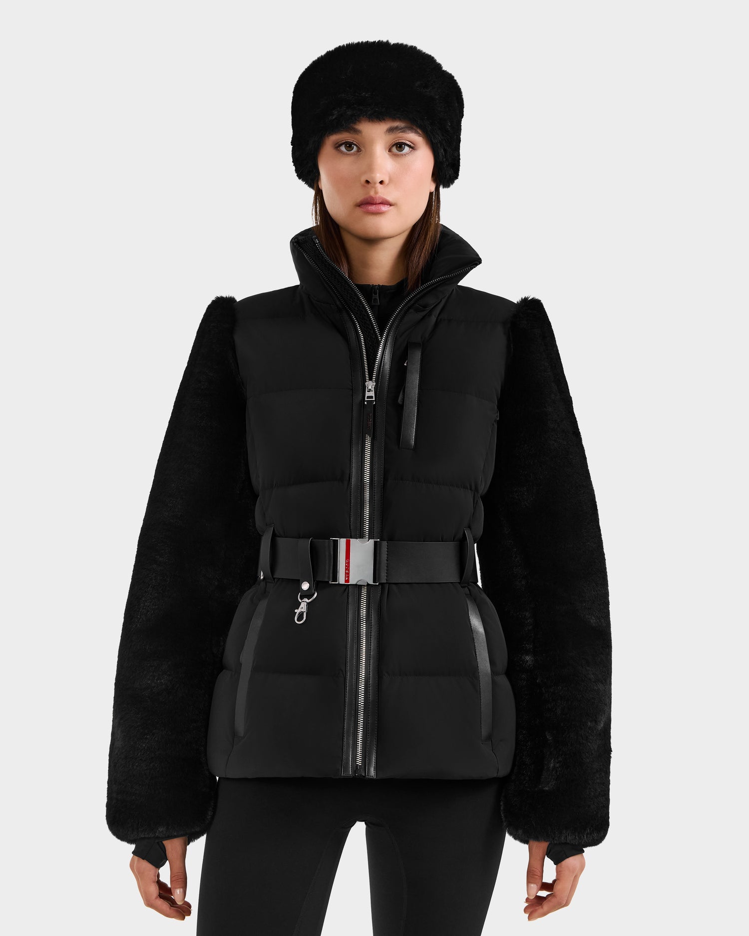 A woman stands facing forward against a plain light gray background, wearing the Rudsak JOANNA F - 8125593 Black faux-fur ski jacket with a belted waist and black pants.