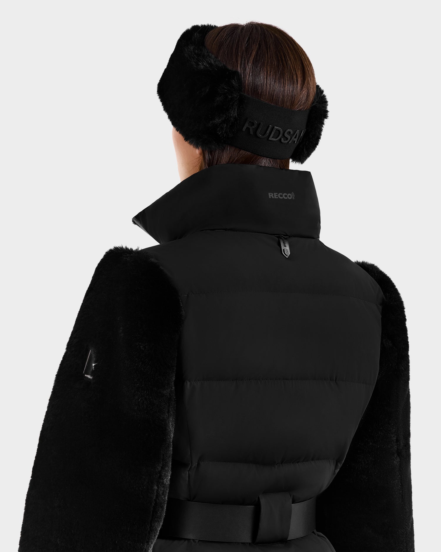 A person shown from behind wears the Rudsak JOANNA F - 8125593 Black women’s down ski jacket with furry sleeves and a matching black furry headband embroidered with RUDSAK, featuring a high collar and RECCO rescue technology label at the back.