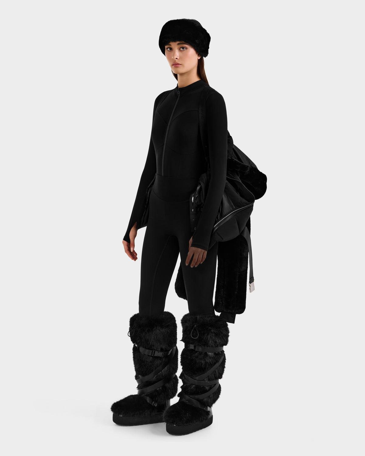 A woman in a black fitted bodysuit, black furry boots, and a black fur hat holds the Rudsak JOANNA F - 8125593 Black women’s down ski jacket draped over her arm against a plain white background.