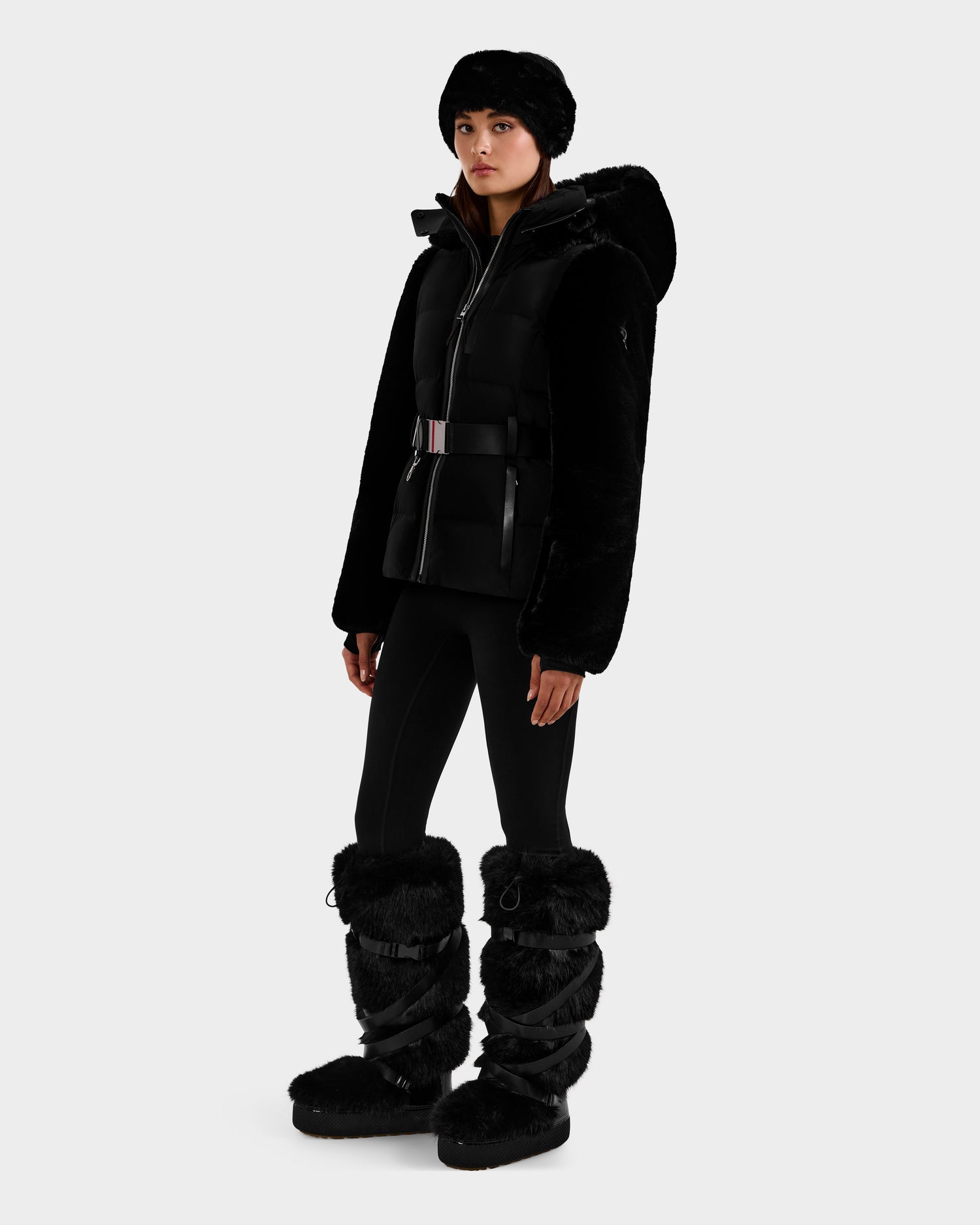A woman wearing the Rudsak JOANNA F - 8125593 Black belted down ski jacket with hood, black fur hat, leggings, and tall furry boots stands against a plain white background.