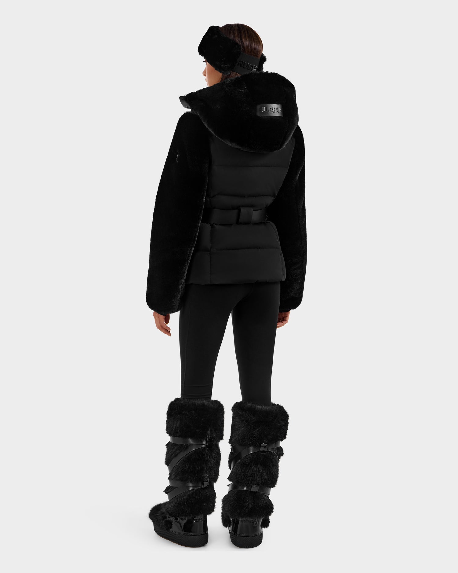 A person faces away, wearing the Rudsak JOANNA F - 8125593 Black faux-fur ski jacket with hood, black headband, fitted pants, and tall fur boots. The winter-ready look is set against a plain white background.