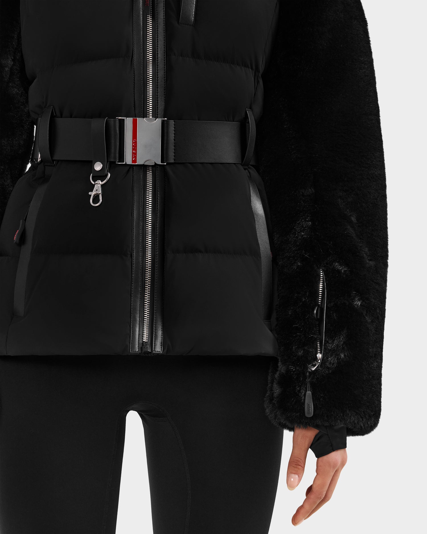A person wears the Rudsak JOANNA F - 8125593 Black women’s down ski jacket with faux fur sleeves and a belted waist, paired with black leggings—shown from shoulders to thighs against a plain light background.