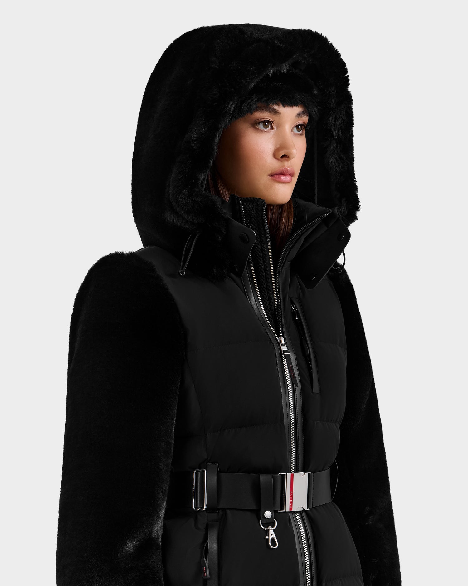 A woman wearing the Rudsak JOANNA F - 8125593 Black down ski jacket with a furry trim, belted waist, and RECCO rescue tech stands against a plain background, looking slightly to the side.