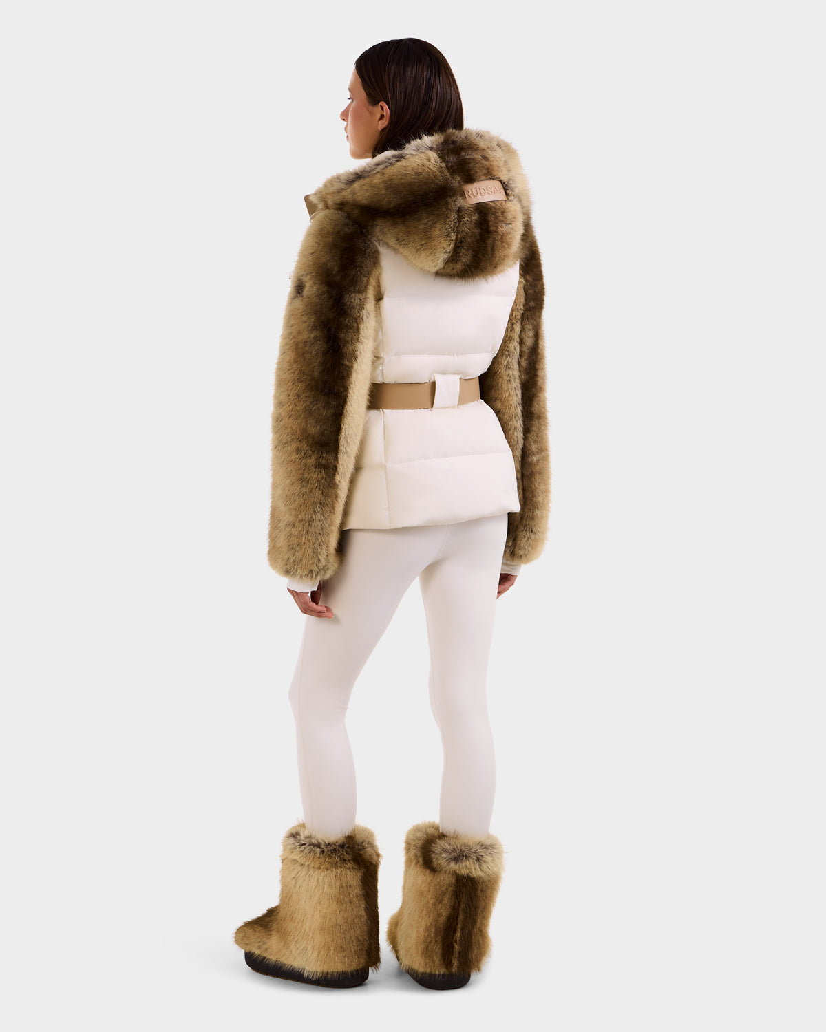 A person faces away, wearing the Rudsak JOANNA F - 8125593 Mocha women's down ski jacket with faux fur trim, fur boots, a beige belt, and white fitted pants against a plain light background.