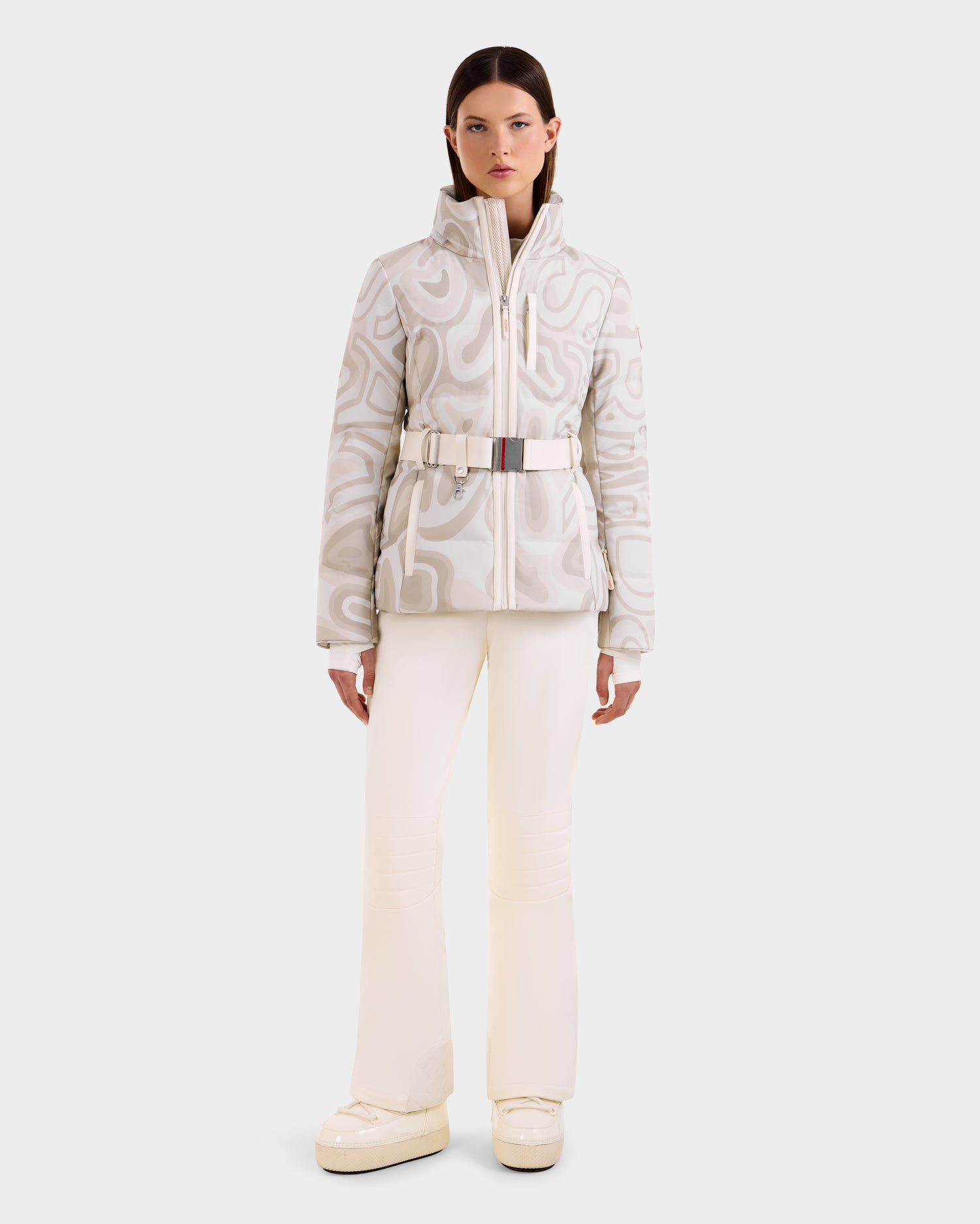 A woman faces forward wearing the Rudsak JOANNA P - 8125623 Vanilla Deja vu belted, patterned down ski jacket with RECCO® rescue tech, paired with ivory flared pants and chunky white shoes against a plain light background.