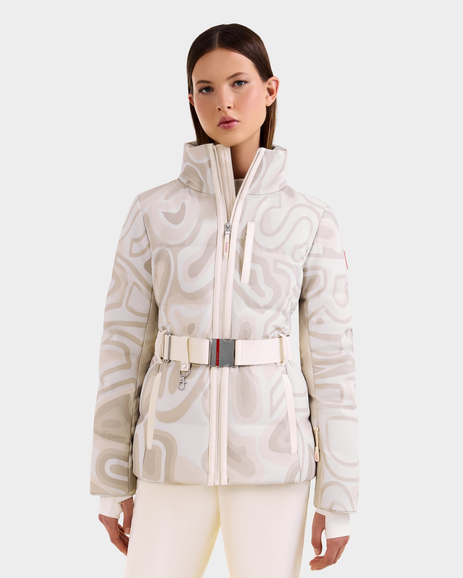 A woman stands facing forward against a light background, wearing the Rudsak JOANNA P - 8125623 Vanilla Deja vu patterned women’s down ski jacket with high collar and belt, paired with white pants featuring RECCO® rescue technology.