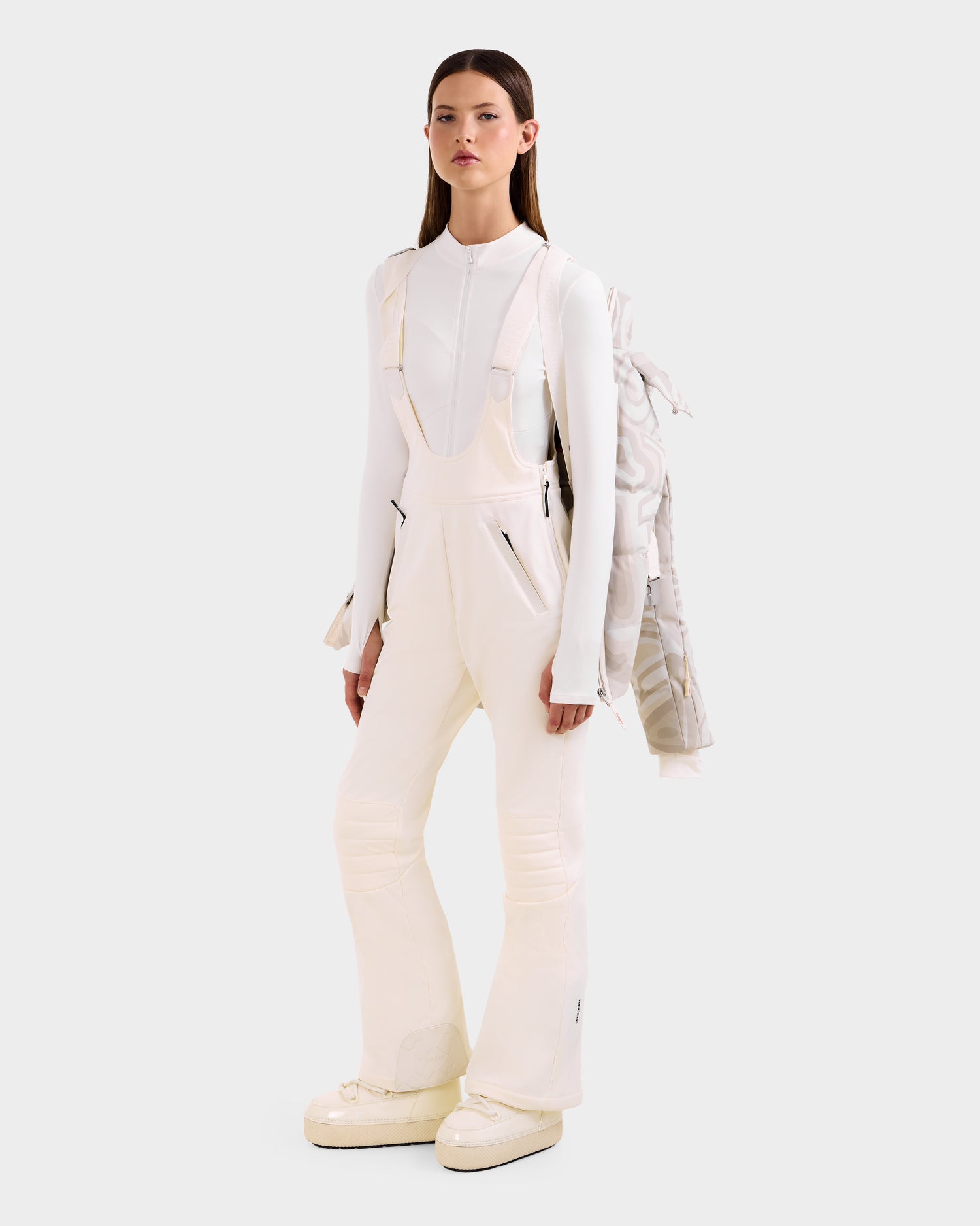 A woman stands against a plain background in a fitted white long-sleeve top, overalls, and platform shoes, with the Rudsak JOANNA P - 8125623 Vanilla Deja vu women's down ski jacket draped over one shoulder.