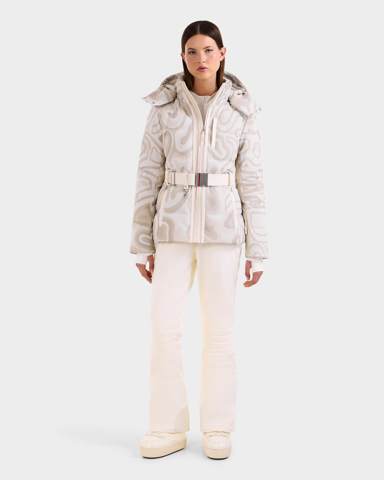 A woman faces forward in the Rudsak JOANNA P - 8125623 Vanilla Deja vu patterned winter jacket with a belt, white pants, and platform boots, against a light background. Pre-order now; ships by November 30th.