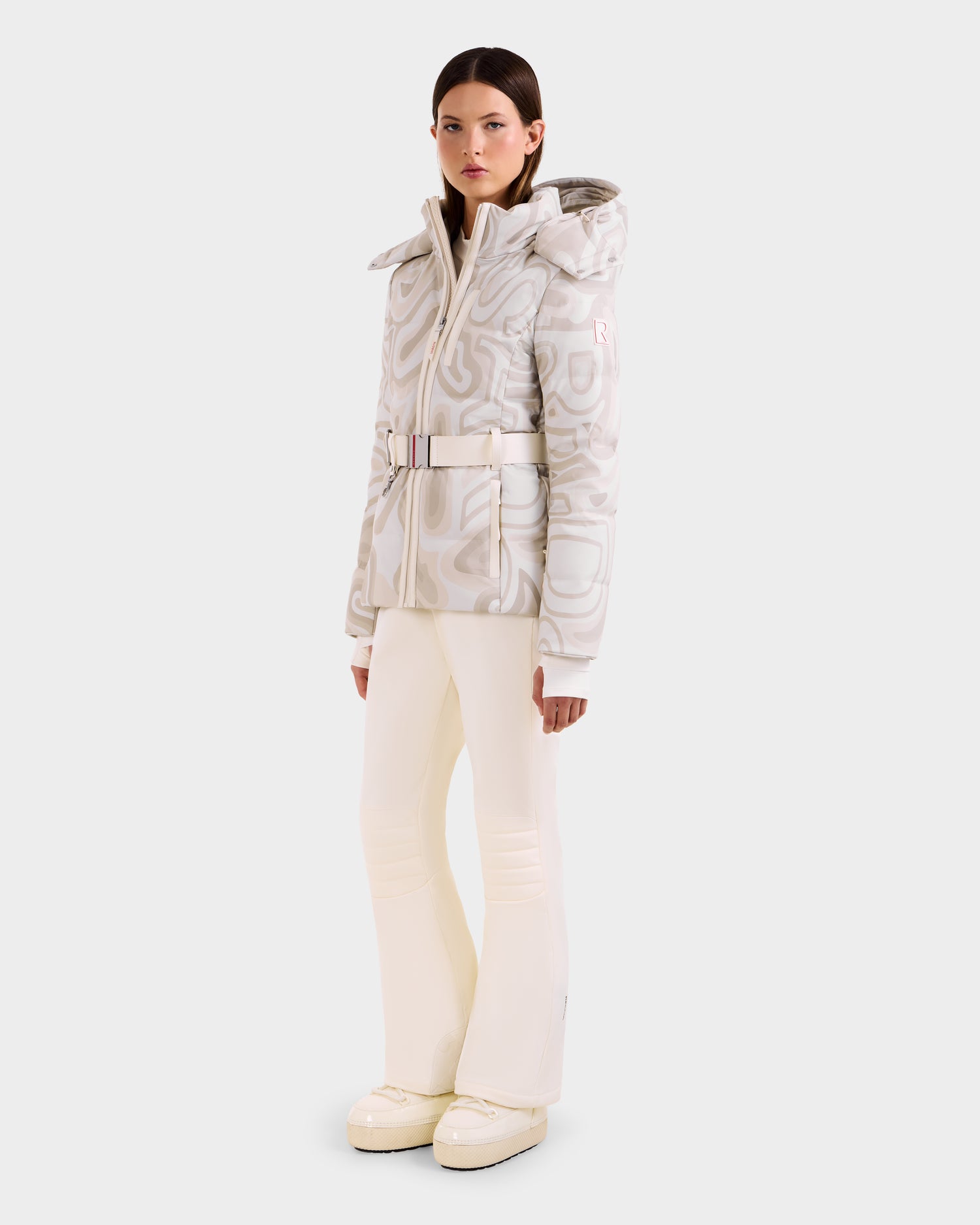 A woman wears the Rudsak JOANNA P - 8125623 Vanilla Deja vu, a cream and white belted women’s down ski jacket with hood featuring RECCO® rescue technology, paired with matching flared pants and snow boots against a light background.