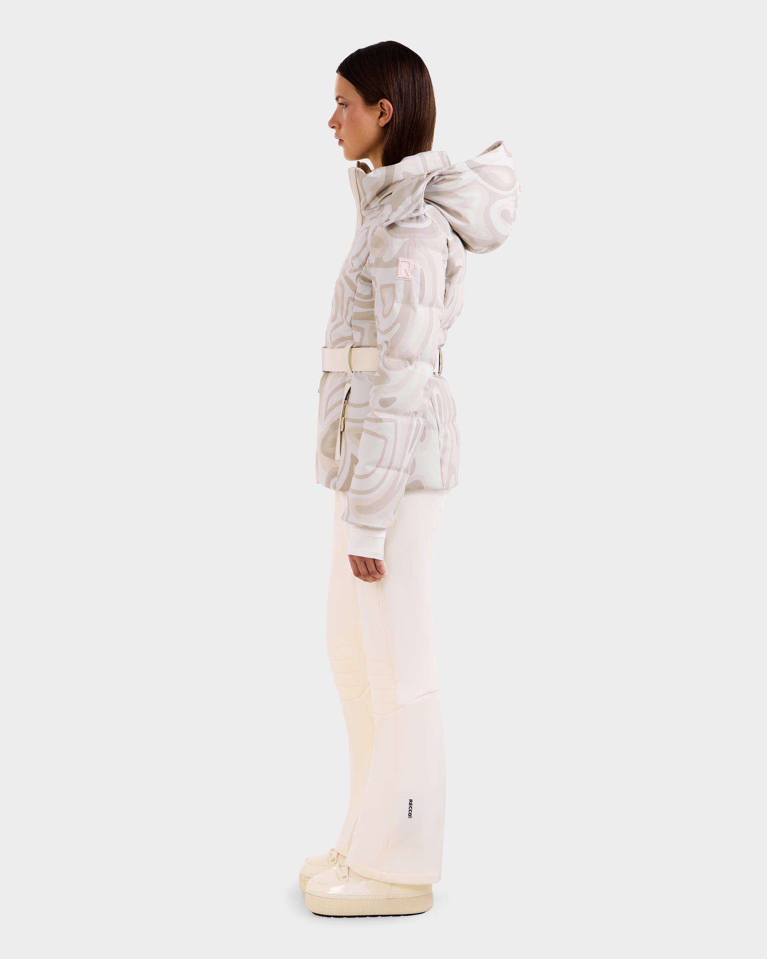 A woman stands in profile wearing the Rudsak JOANNA P - 8125623 Vanilla Deja vu patterned women’s down ski jacket with RECCO® rescue technology, paired with cream flared pants and matching shoes against a plain white background.