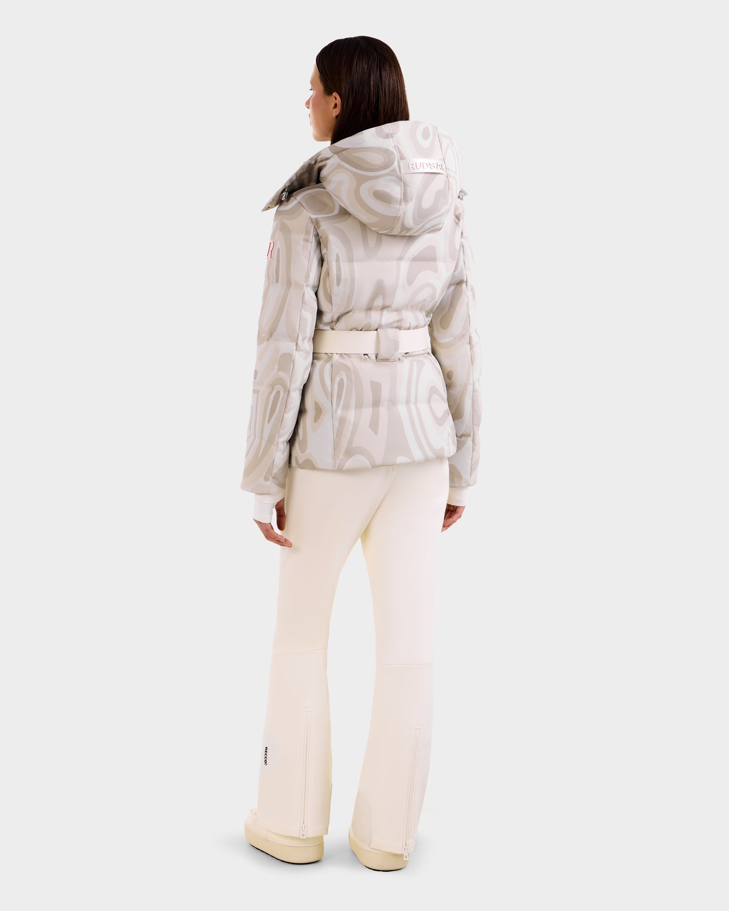 A woman faces away, wearing the Rudsak JOANNA P - 8125623 Vanilla Deja vu women’s patterned light beige down ski jacket with RECCO® rescue tech, hood, and belt, paired with cream pants and beige shoes against a plain white background.
