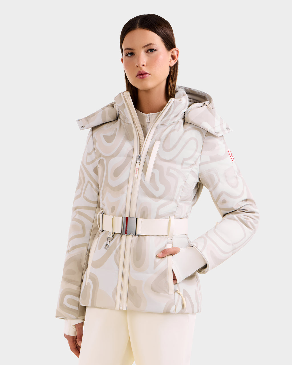 A woman wears the Rudsak JOANNA P - 8125623 Vanilla Deja vu fitted women's down ski jacket with a belted waist, hood, and high collar, featuring premium insulation, standing with one hand in her pocket against a plain background.