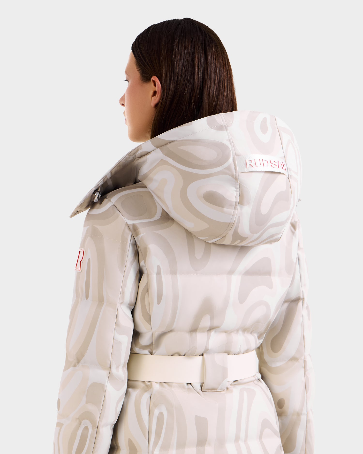 A person with straight brown hair wears the Rudsak JOANNA P - 8125623 Vanilla Deja vu women's down ski jacket, featuring a hood with "RUDSAK" and RECCO® rescue technology, seen patterned and belted from the back and side.