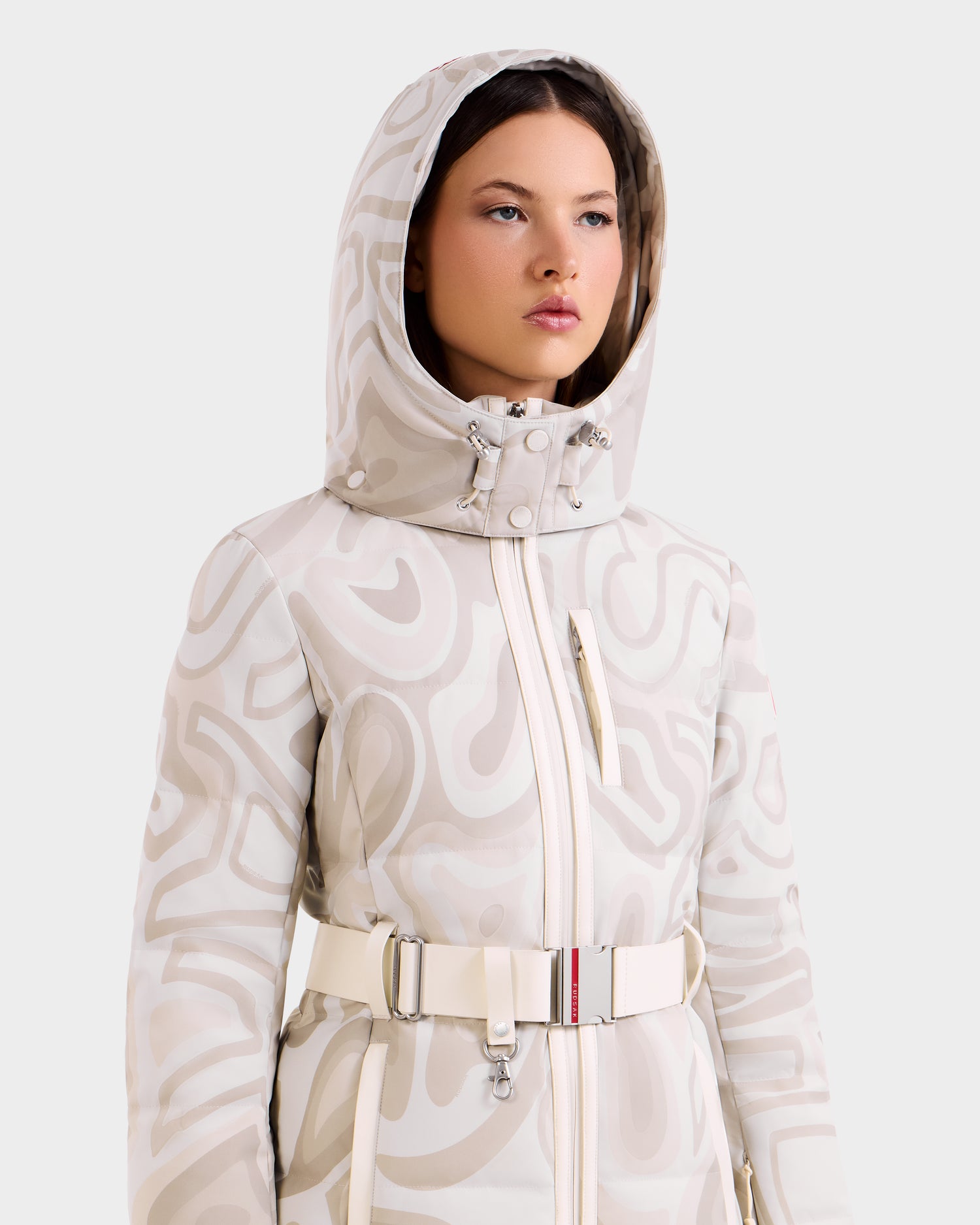 A woman wearing the Rudsak JOANNA P - 8125623 Vanilla Deja vu women’s down ski jacket with RECCO® rescue technology and a belted high collar stands against a plain background, gazing slightly upward with a neutral expression.