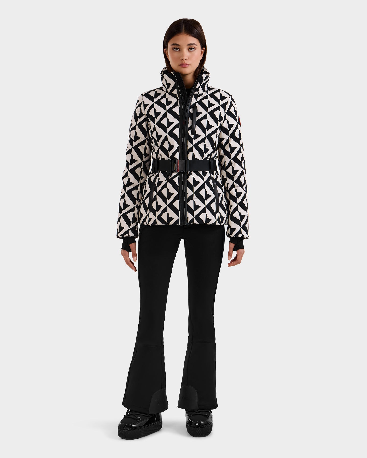 A woman faces forward, wearing the Rudsak JOANNA P - 8125623 Vanilla Monogram down ski jacket with a belt, paired with black flared pants and black shoes, standing against a plain white background.