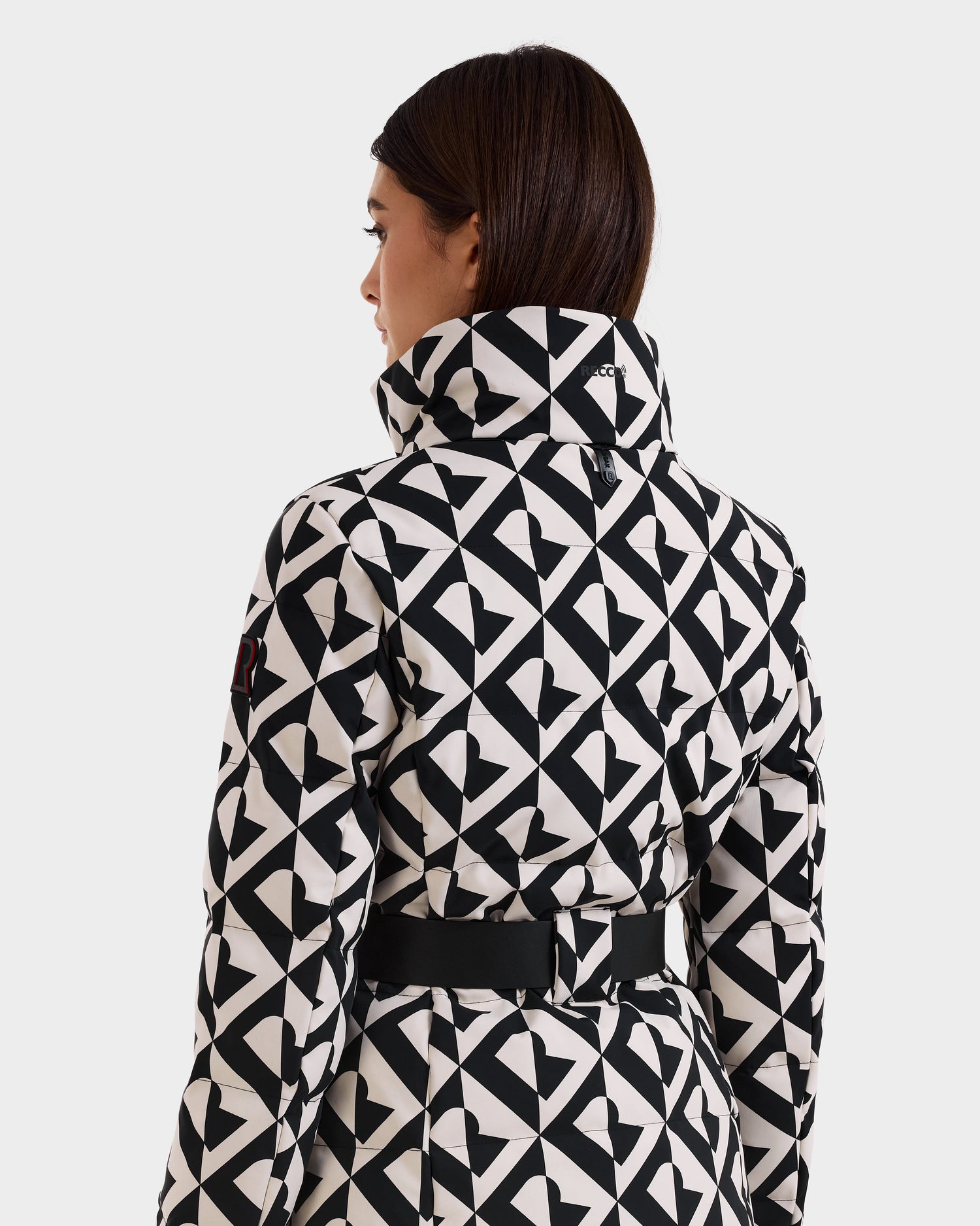 A woman with straight brown hair is shown from the back, wearing the Rudsak JOANNA P - 8125623 Vanilla Monogram women’s down ski jacket, featuring a high collar, black waist belt, and a discreet ski-pass pocket.
