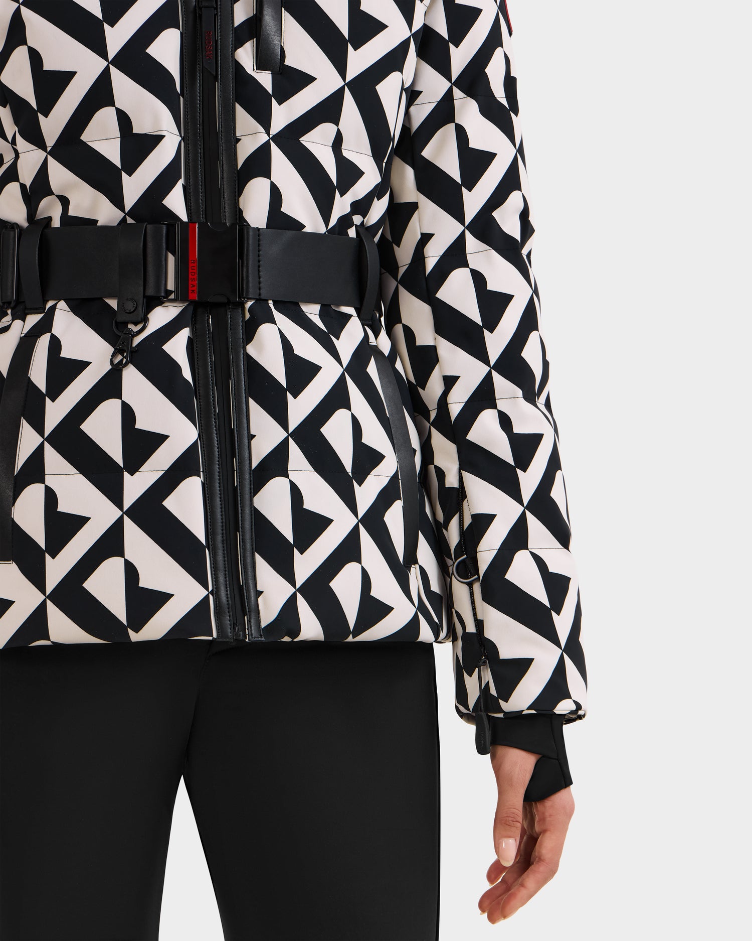A person wearing the Rudsak JOANNA P - 8125623 Vanilla Monogram women’s down ski jacket with a black and white geometric pattern, heart and triangle shapes, a belt, and black pants. Only the lower half is visible.