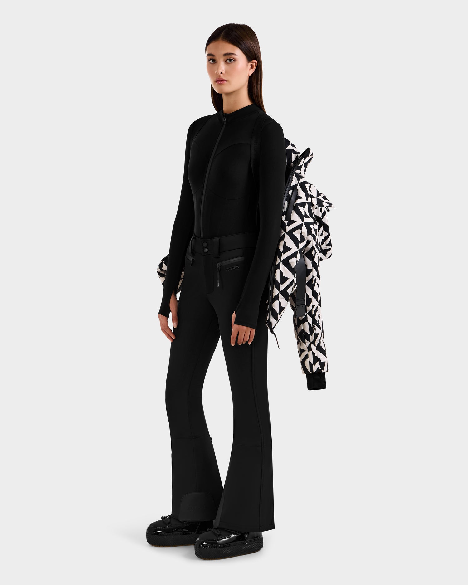 A woman poses against a plain background wearing a fitted black zip-up top, flared pants, and boots. The Rudsak JOANNA P - 8125623 Vanilla Monogram women’s down ski jacket with a ski-pass pocket is draped over one shoulder.