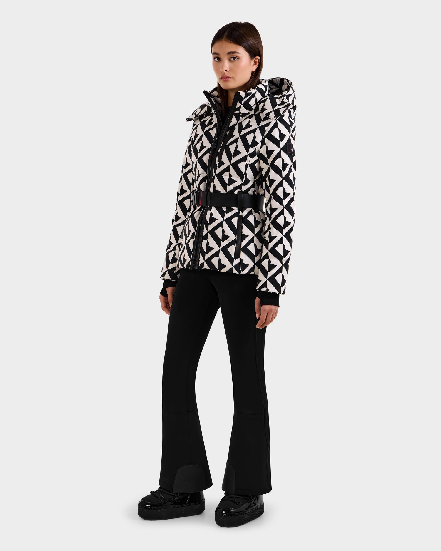 A woman with straight dark hair poses against a white background wearing the Rudsak JOANNA P - 8125623 Vanilla Monogram down ski jacket with a geometric pattern, black flared pants, and black shoes.